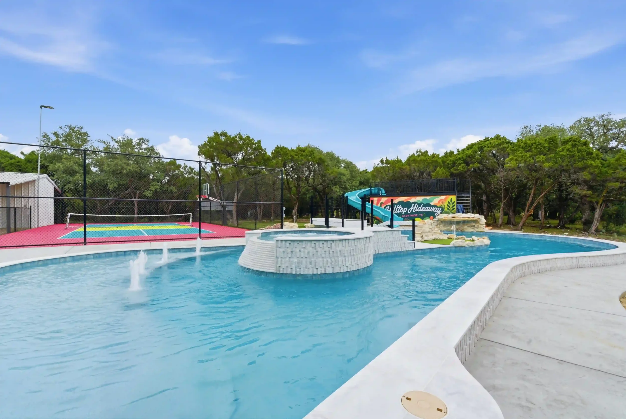 A water park with a large pool, a colorful slide, a smaller splash pad with a basketball hoop, and a sign that reads 'Hilltop Hideaway' surrounded by trees under a blue sky.