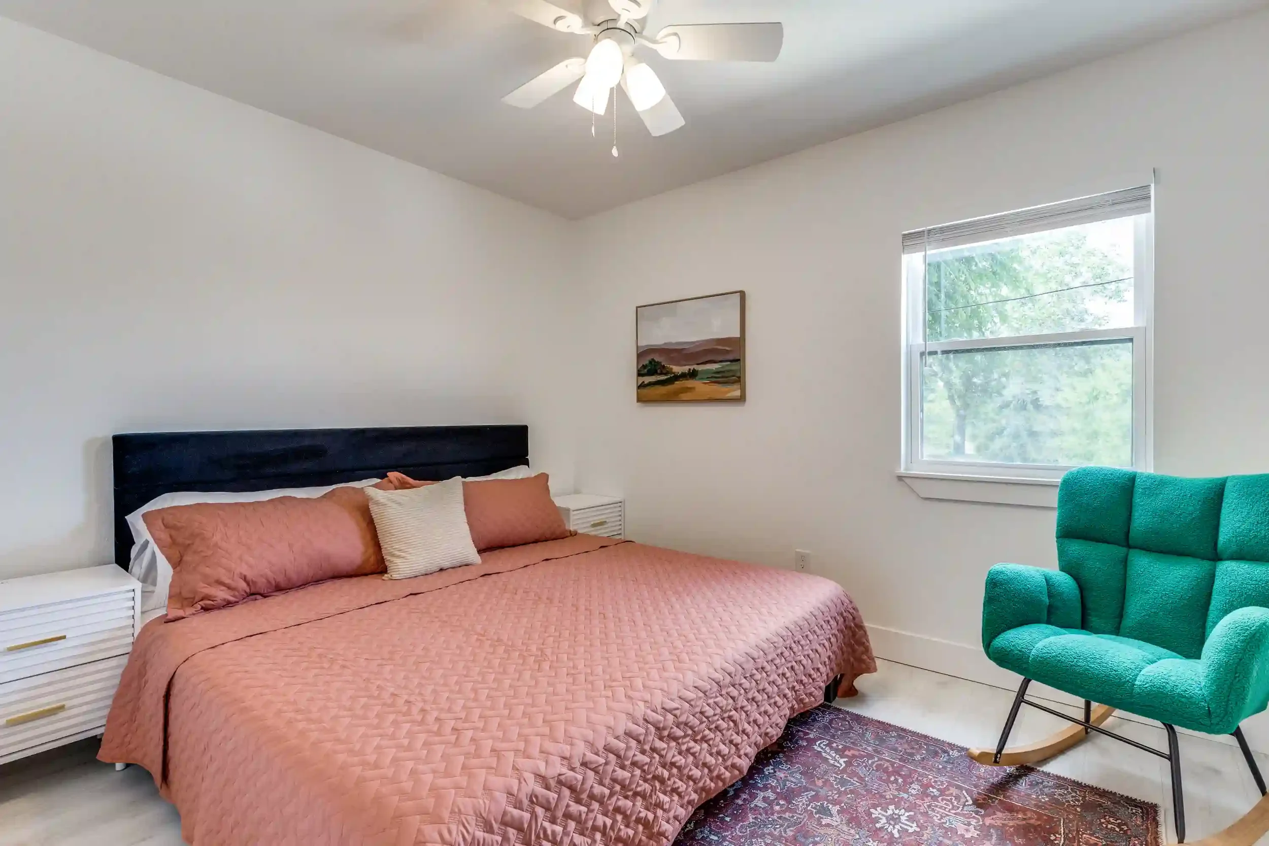 A bedroom with a bed covered in a pink quilt, white pillows, and a black headboard. There is a white nightstand with gold handles beside the bed. A colorful painting hangs on the white wall above the bed. A teal rocking chair is in the corner near a 