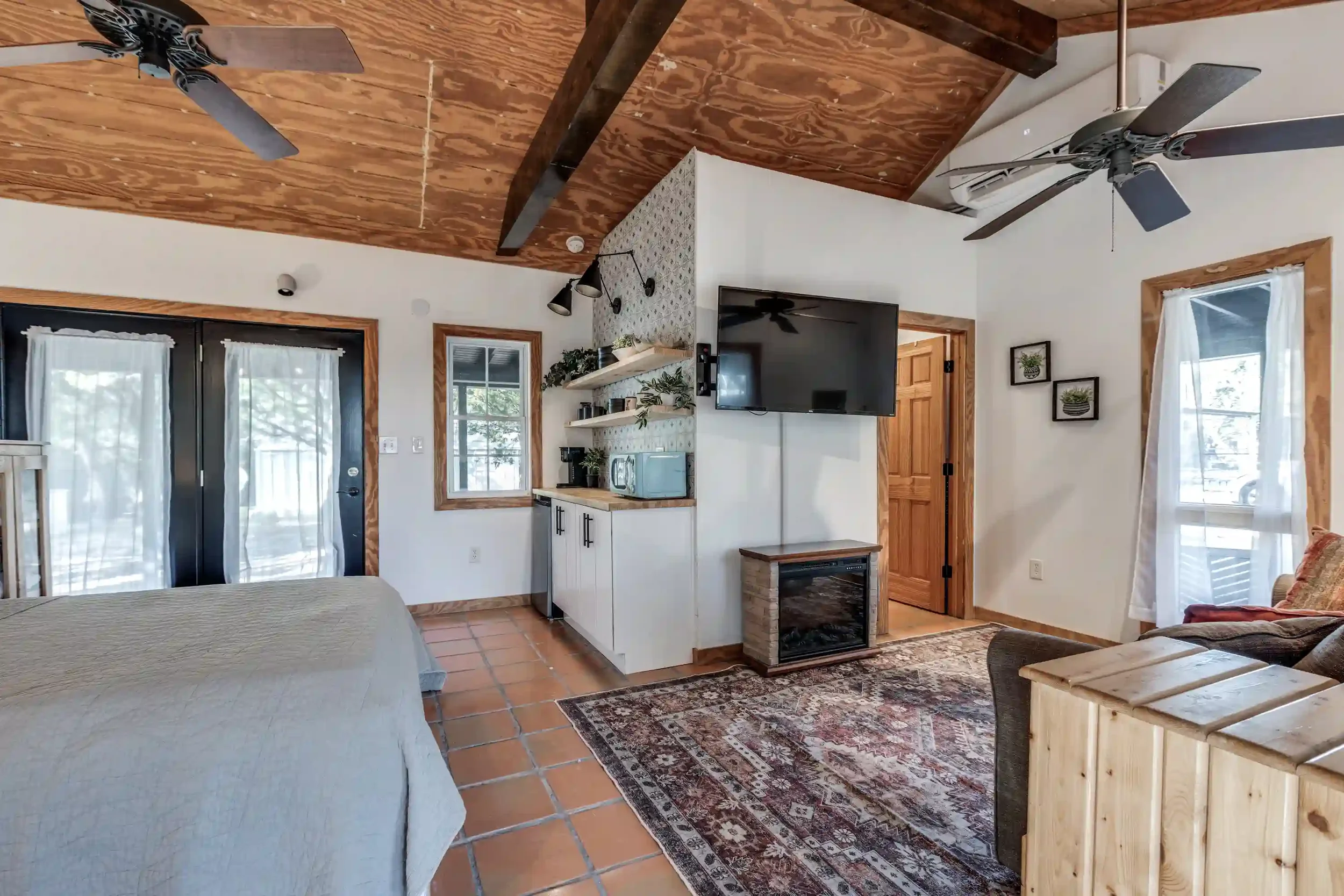 Living room with wooden ceiling, ceiling fans, white walls, windows with white curtains, a large flat-screen TV mounted on the wall, a wooden door, and a sitting area with a patterned rug and a chair.