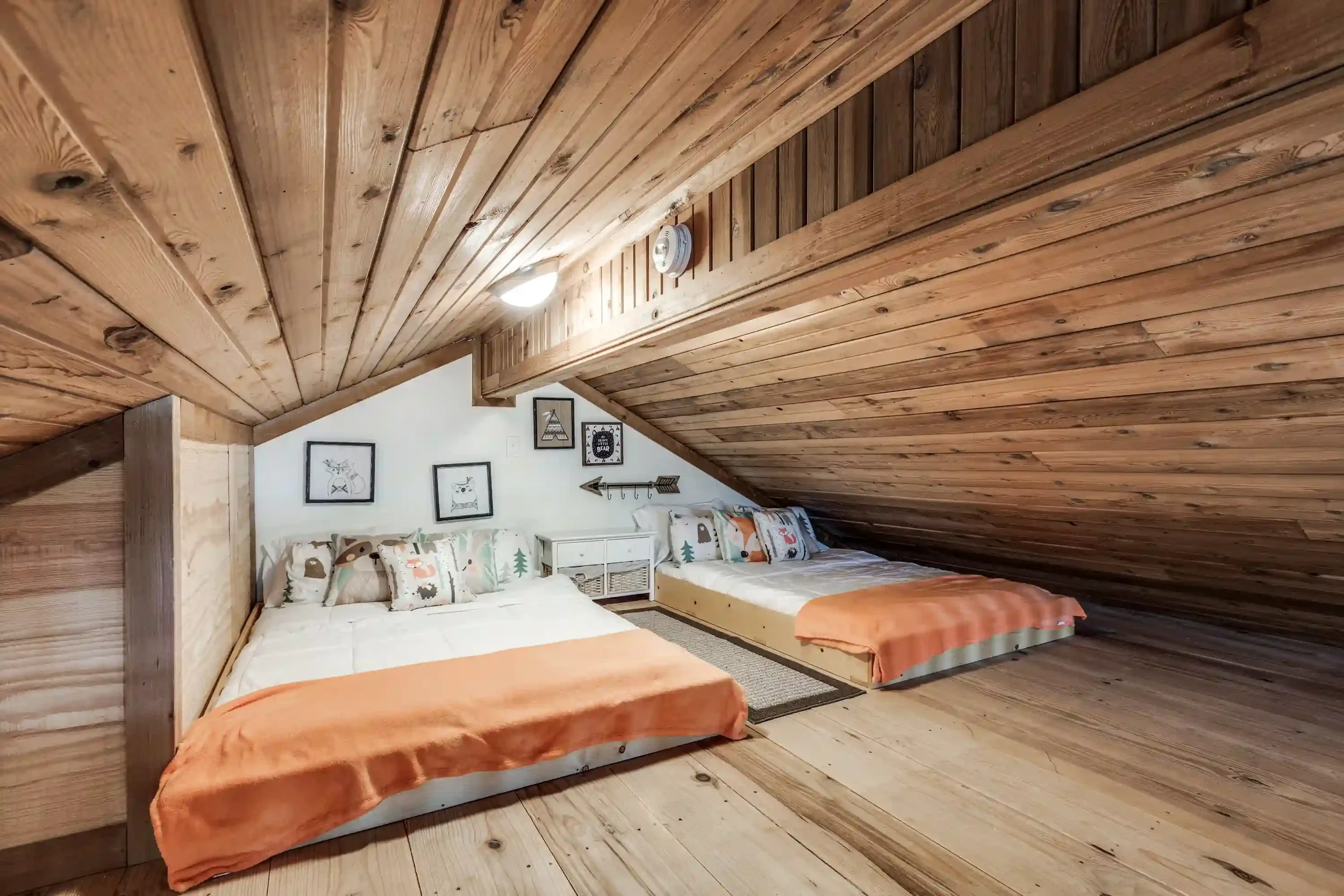 A bedroom with a wooden sloped ceiling and wall, decorated with framed pictures of animals and forest scenes. There are two beds with white bedding and patterned pillows, and a small white nightstand between them.