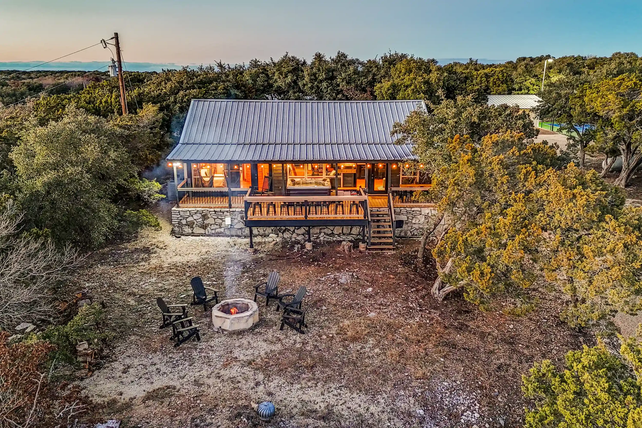 A house with a metal roof, large porch, and outdoor seating area, surrounded by trees and a yard with a fire pit and chairs, during sunset.