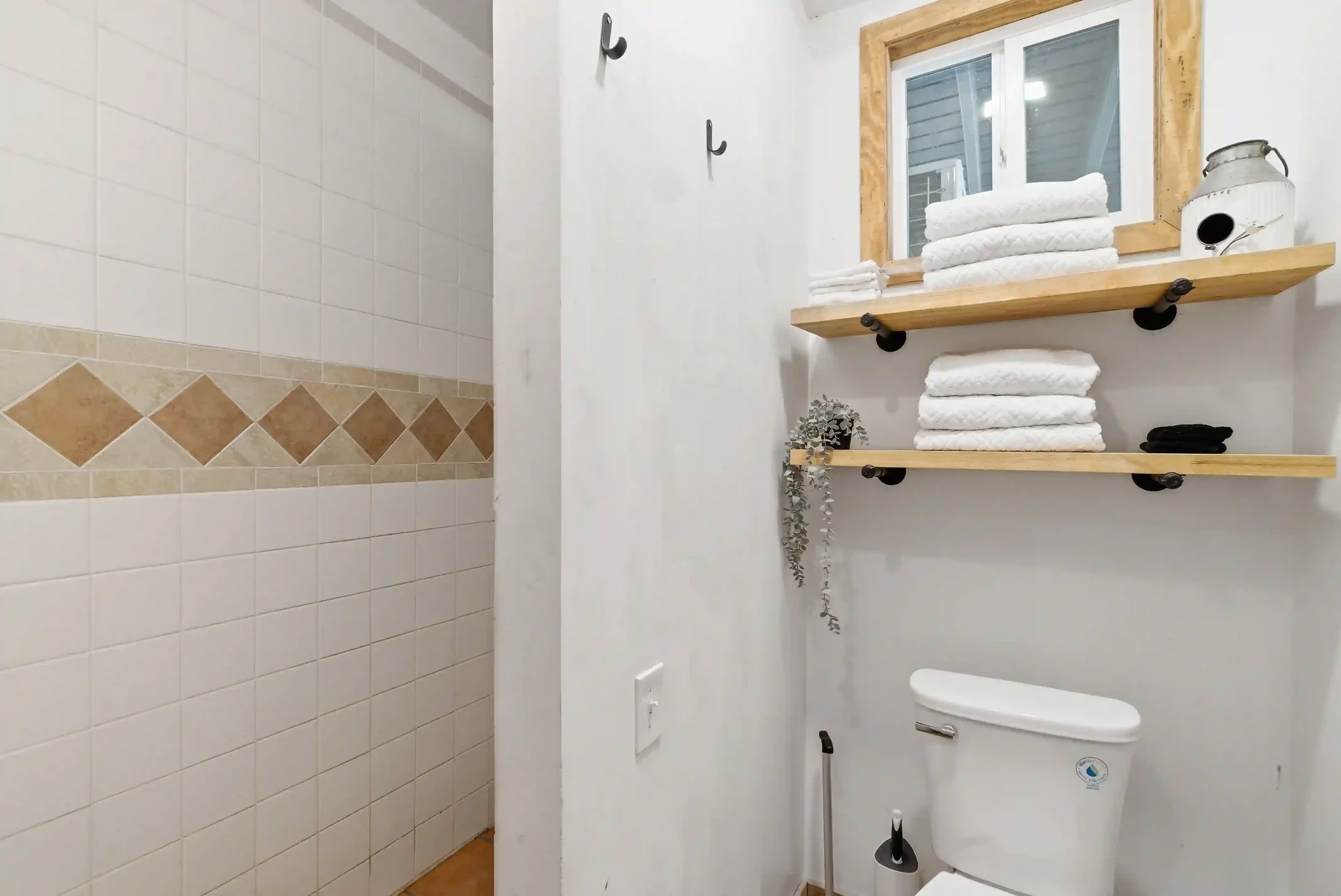 Bathroom with white tiled walls and beige tile accents on the shower wall. A wooden shelf holds white towels and a decorative jar. A window with a wooden frame looks outside. A white toilet is visible.
