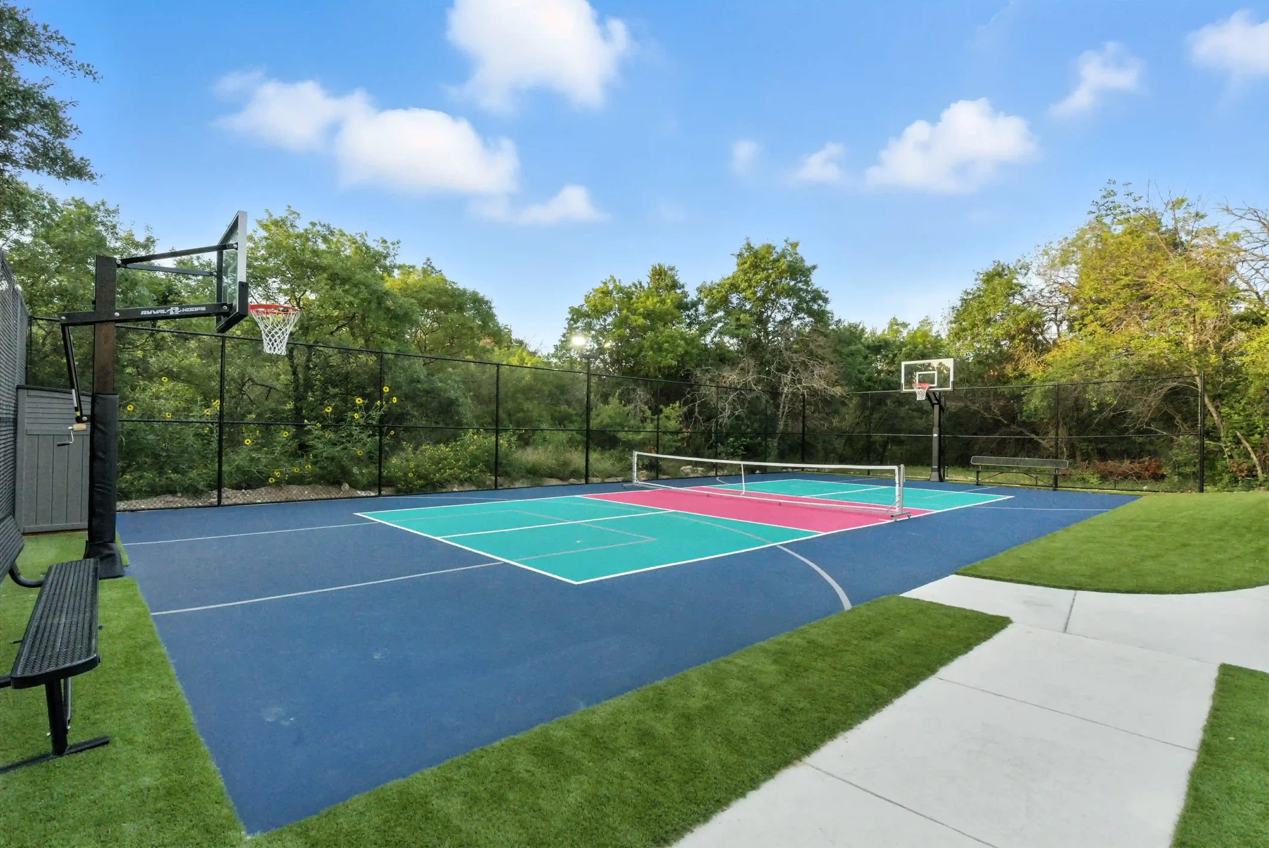 A colorful outdoor sports court with basketball hoops, a net for pickleball or tennis, surrounded by green trees, and a blue sky with a few clouds.