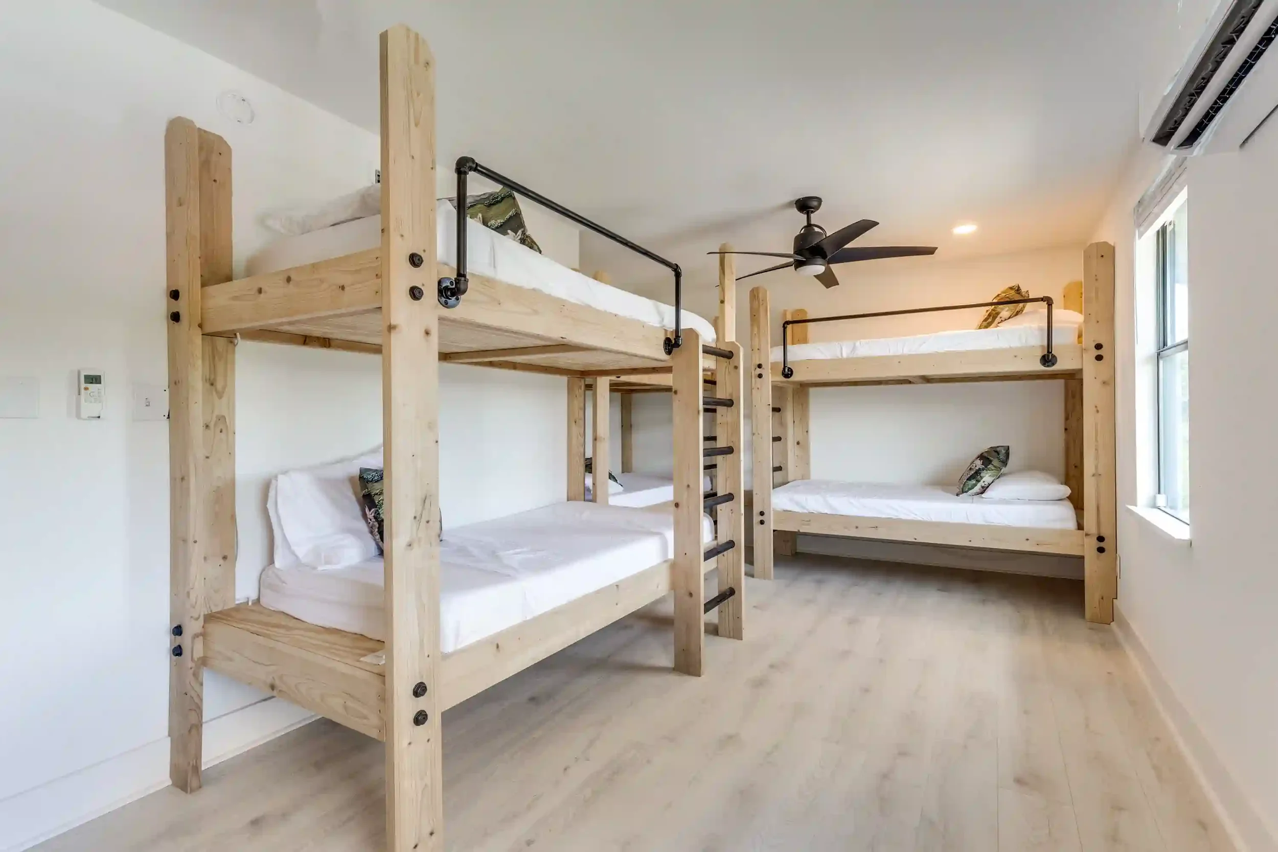 A room with four bunk beds made of light wood, each with white bedding and a decorative pillow. The room has white walls, a ceiling fan, an air conditioning unit, and a window letting in natural light.