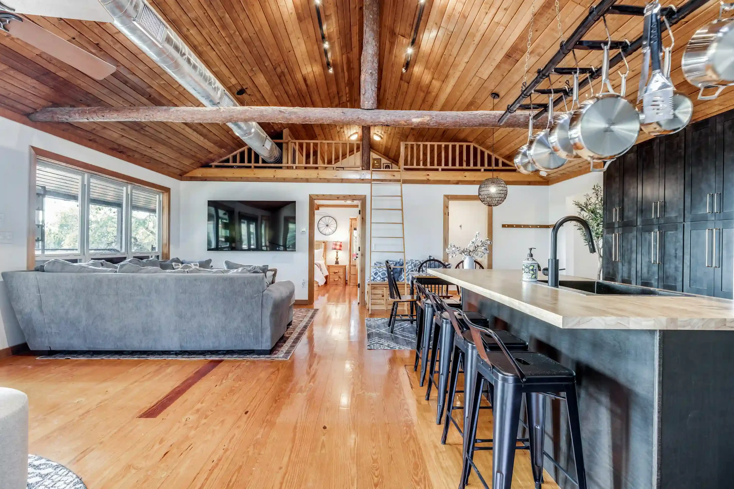 Open-concept living space with wooden vaulted ceiling, exposed beams, large windows, gray sofa, flat-screen TV, and kitchen island with black bar stools.