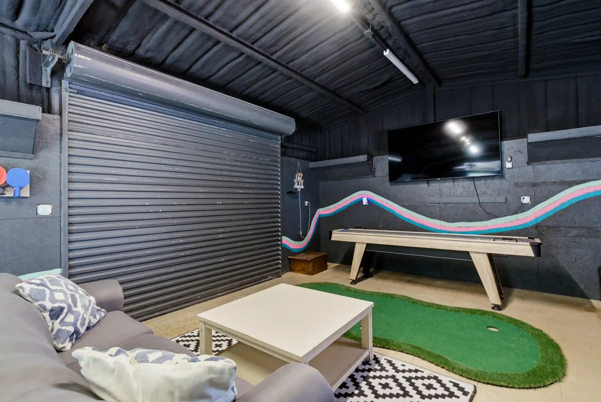 A cozy indoor space featuring a gray sofa with patterned pillows, a white coffee table, a green rug, a mural with rainbow stripes on the black wall, a flat-screen TV, and a black wall with a rainbow-themed design.