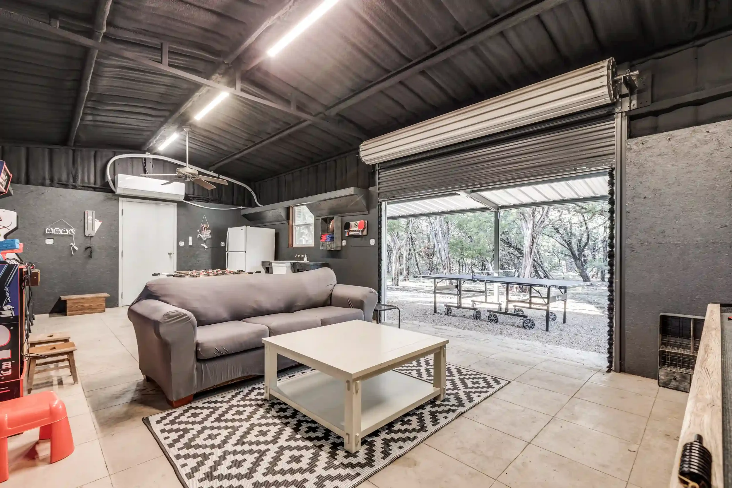 Inside a garage or workshop with a gray couch, white coffee table, black and white patterned rug, and open garage door revealing outdoor trees and patio