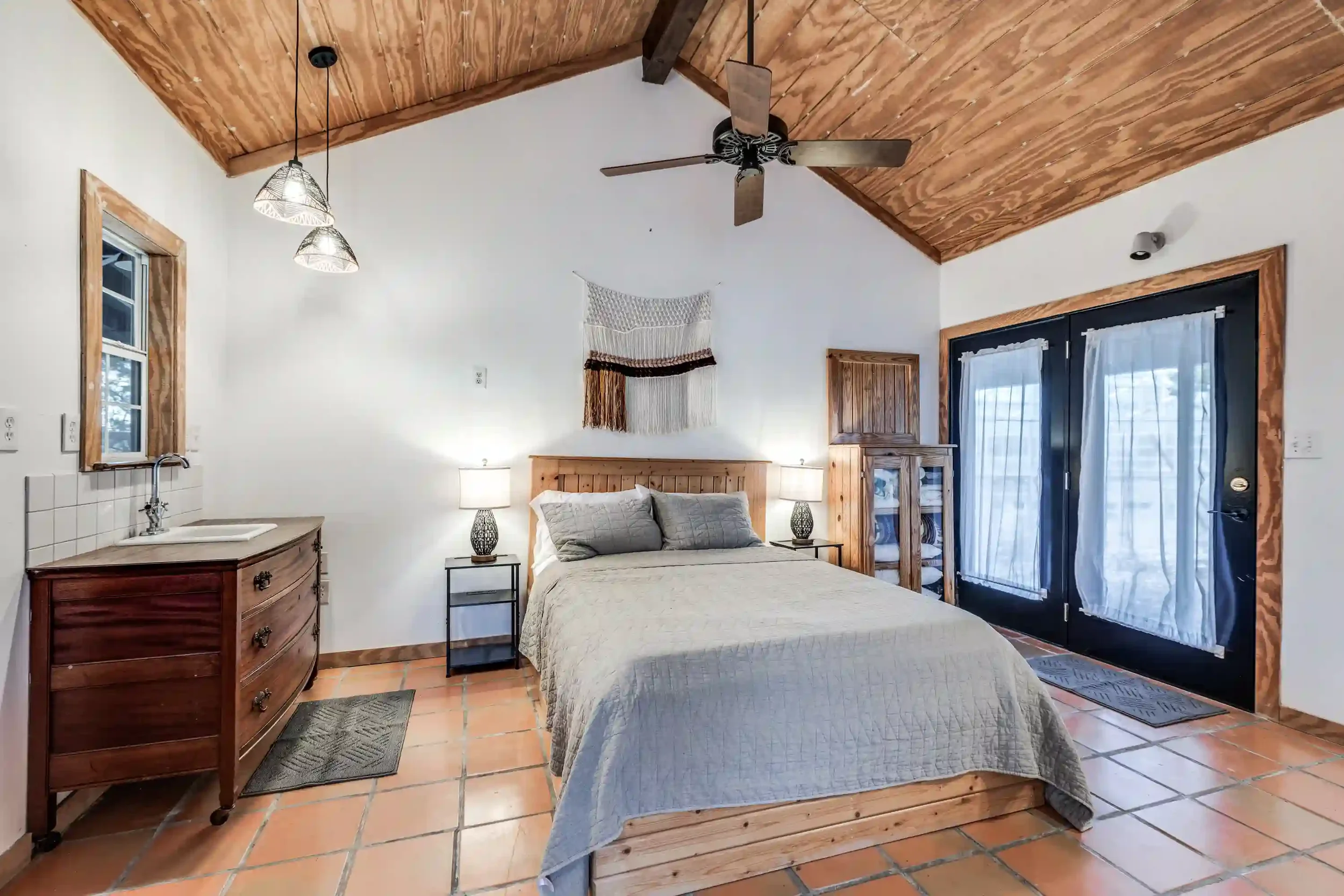 A cozy bedroom with a wooden ceiling and a ceiling fan. The room has a bed with a quilted bedspread, two bedside tables with lamps, a wooden dresser, and large sliding glass doors with white curtains. There is a macramé wall hanging above the bed and