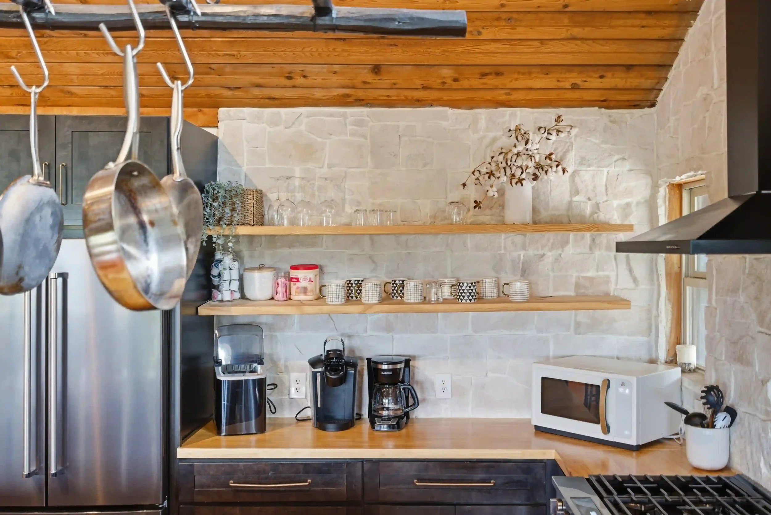 A modern kitchen with a stainless steel refrigerator, wooden open shelves with glassware and decorative items, a coffee maker, microwave, and various kitchen utensils. The wall is made of light-colored stone, and there is a wooden ceiling with a slan