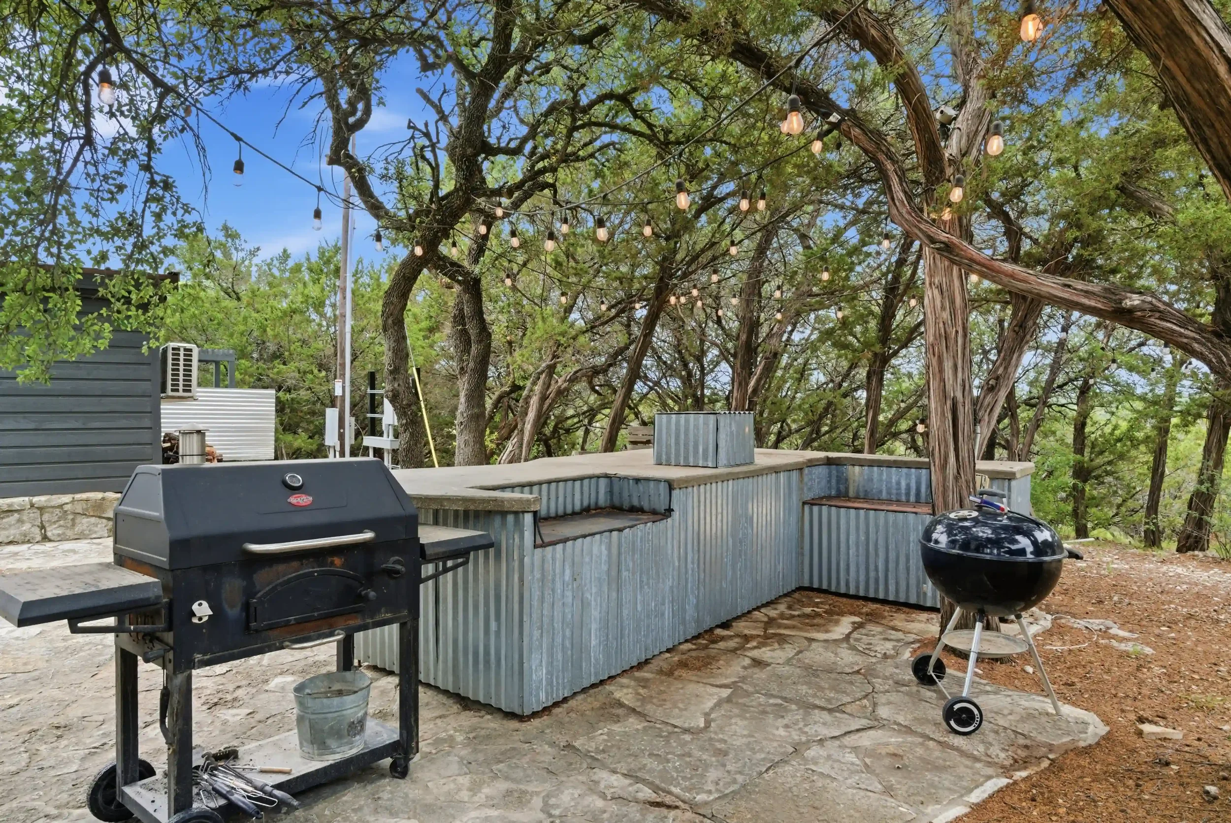 An outdoor patio area with a grill, charcoal barbecue, and a black smoker, surrounded by trees with string lights overhead, and a clear blue sky.