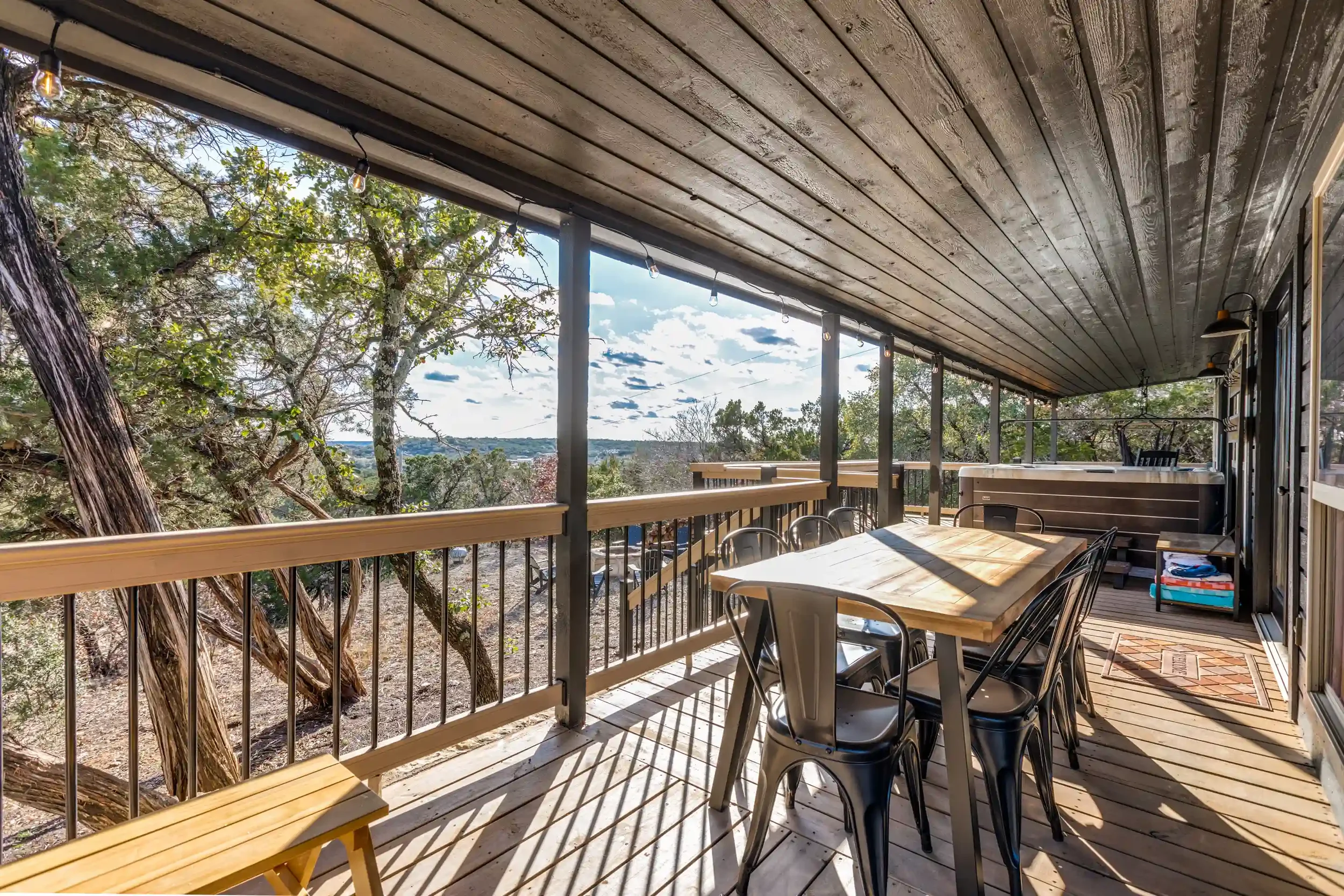 Wood-paneled covered patio with a long outdoor table and chairs, string lights, trees, and a view of the sky and landscape in the background.