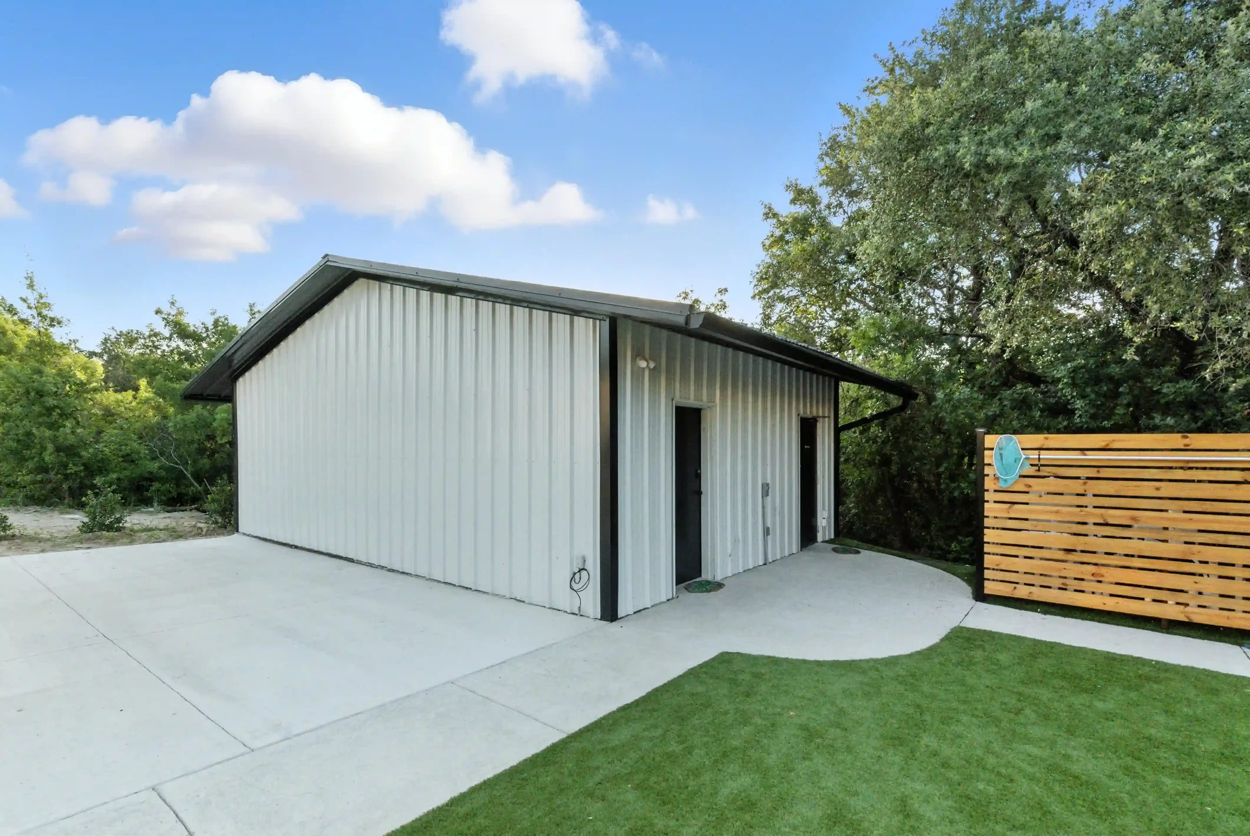 A small metal building with vertical siding, a black door, and a concrete patio in a backyard, surrounded by green trees and a wooden fence with a blue object hanging on it, under a blue sky with white clouds.