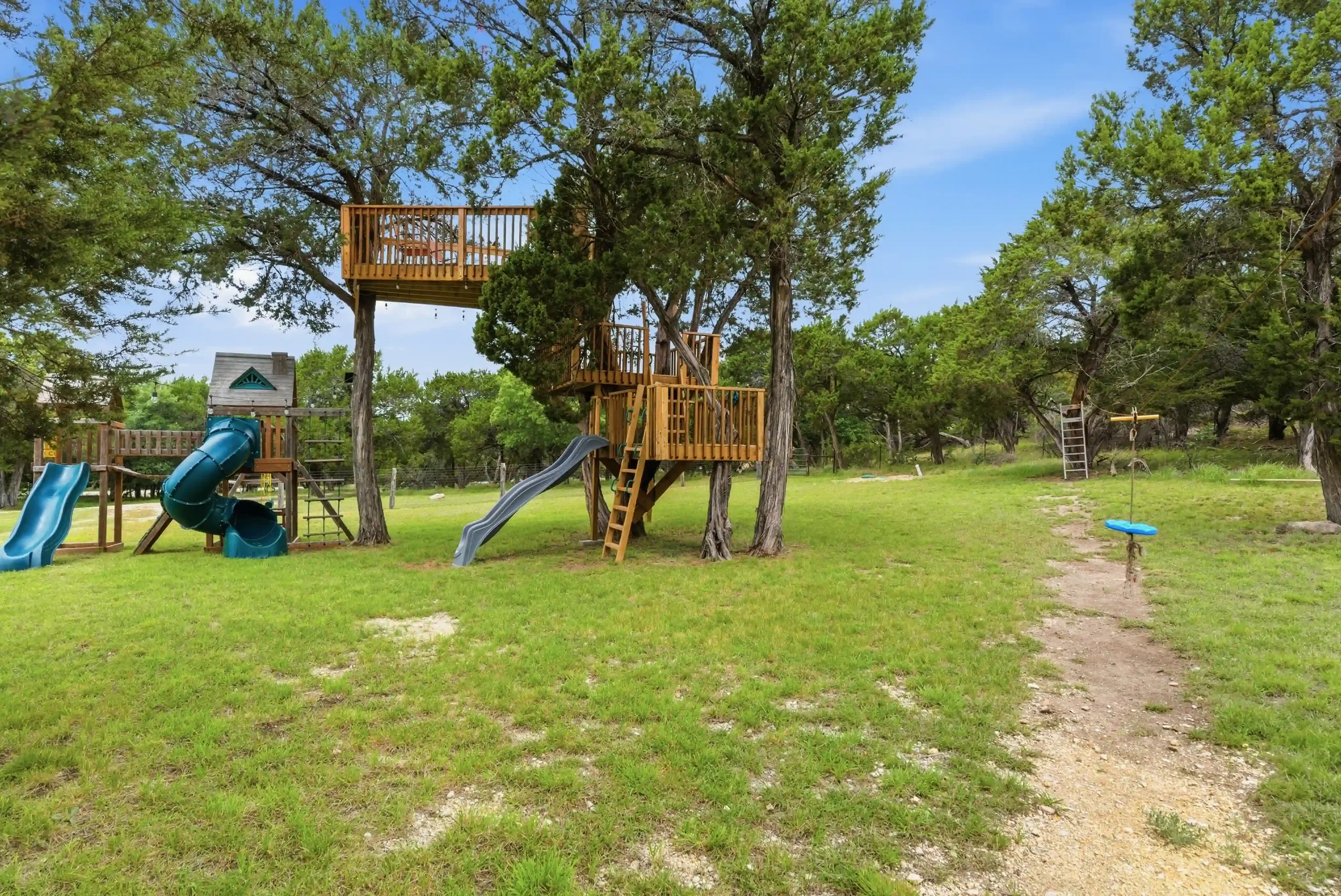 A backyard play area with a wooden treehouse, slides, swings, and lush green trees under a partly cloudy sky.