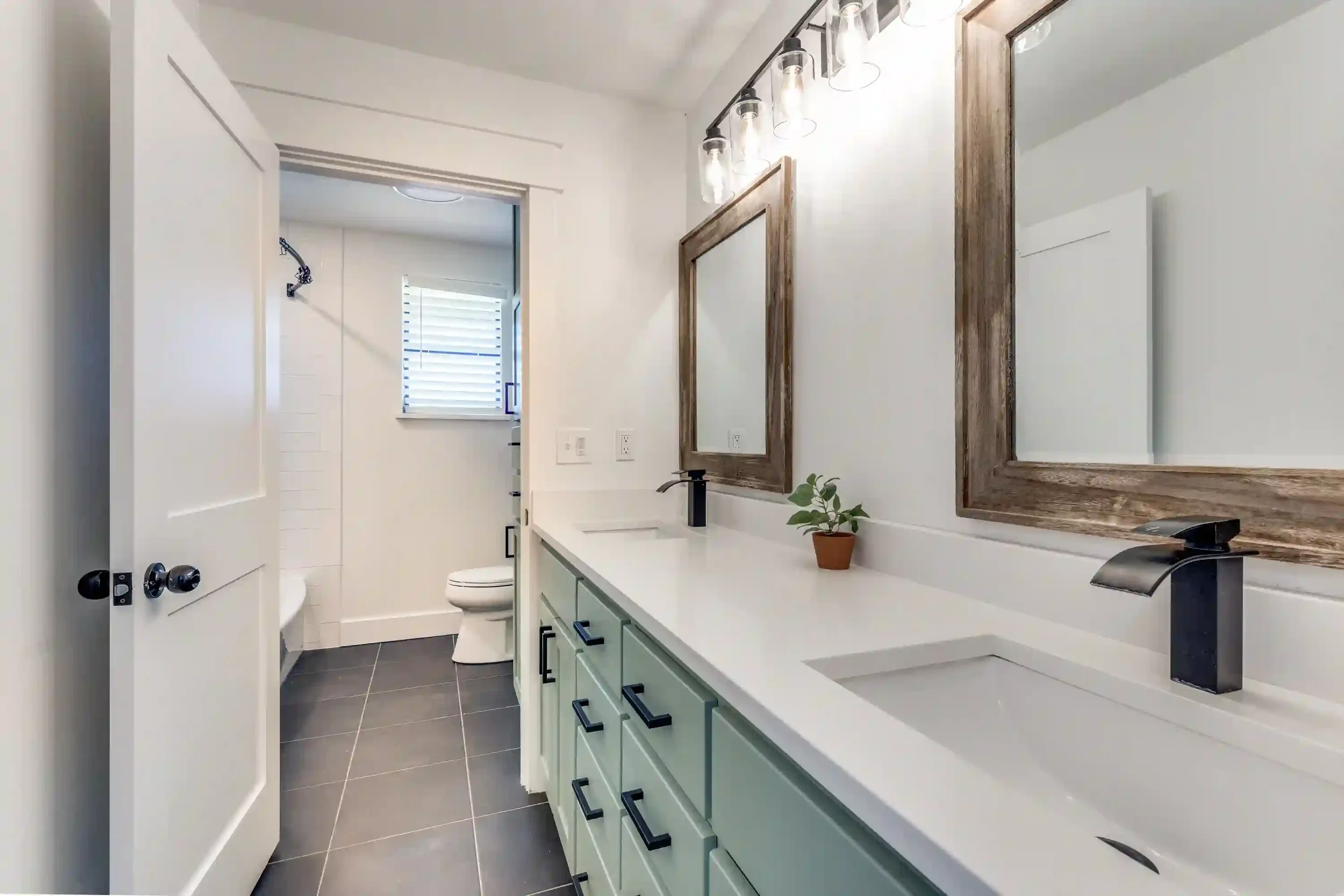 Modern bathroom with white walls, black fixtures, and a double mirror with wooden frames above a white countertop. There is a small potted plant on the counter, and a window with white blinds. The bathroom also features a shower area with a black sho