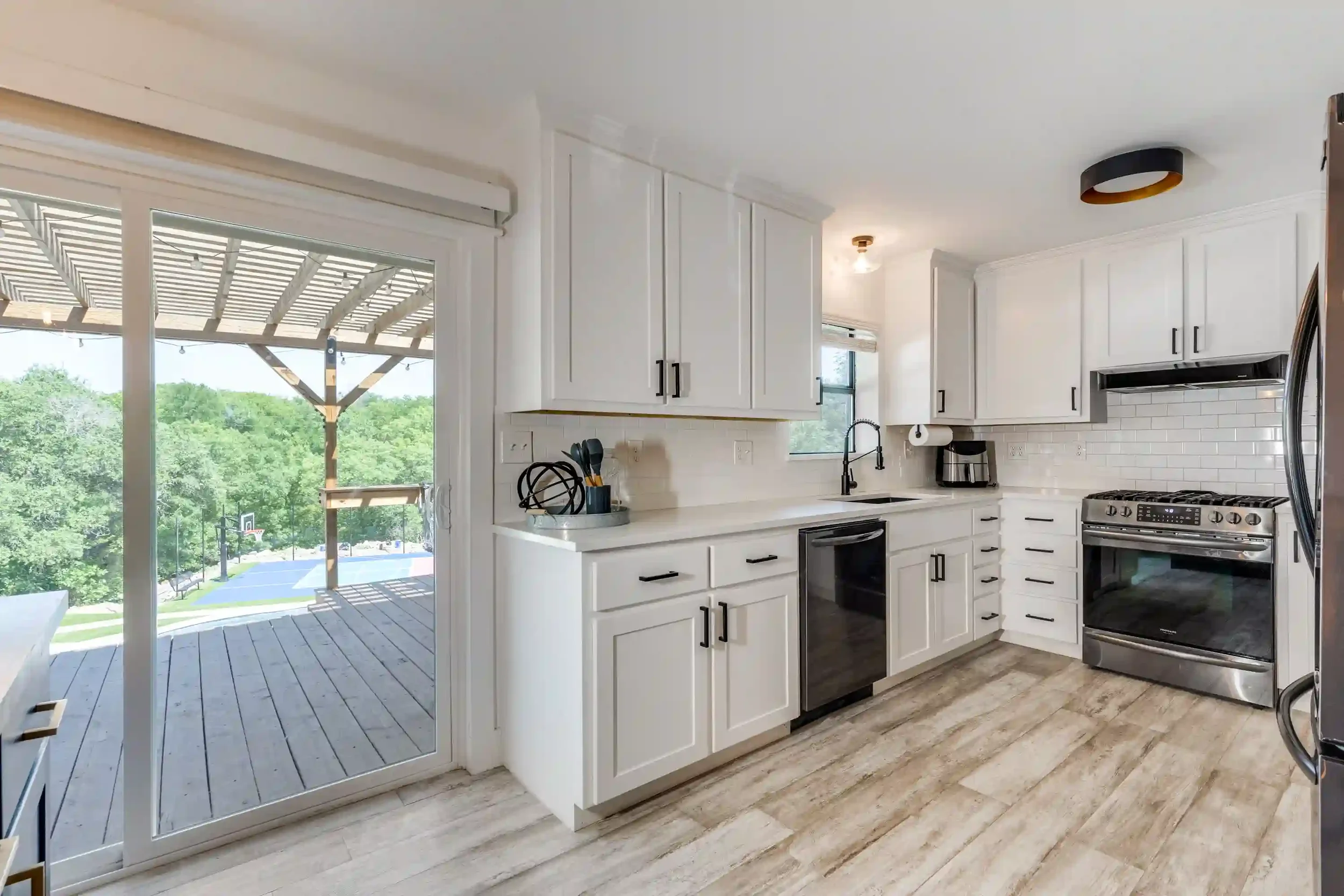 Modern kitchen with white cabinets, stainless steel appliances, and a sliding glass door leading to a deck with trees outside.