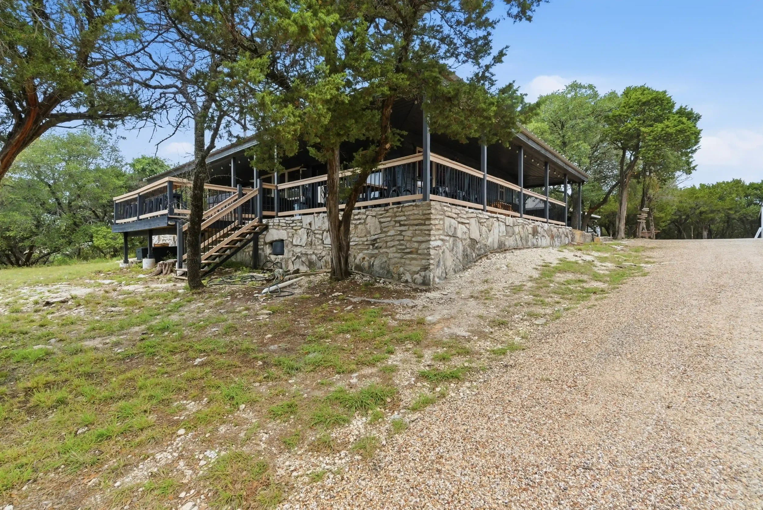A raised wooden deck with a black railing surrounds a house partially obscured by trees. A set of stairs leads up to the deck. The house has a stone foundation, and the area around the house is grassy with some trees and a gravel driveway.