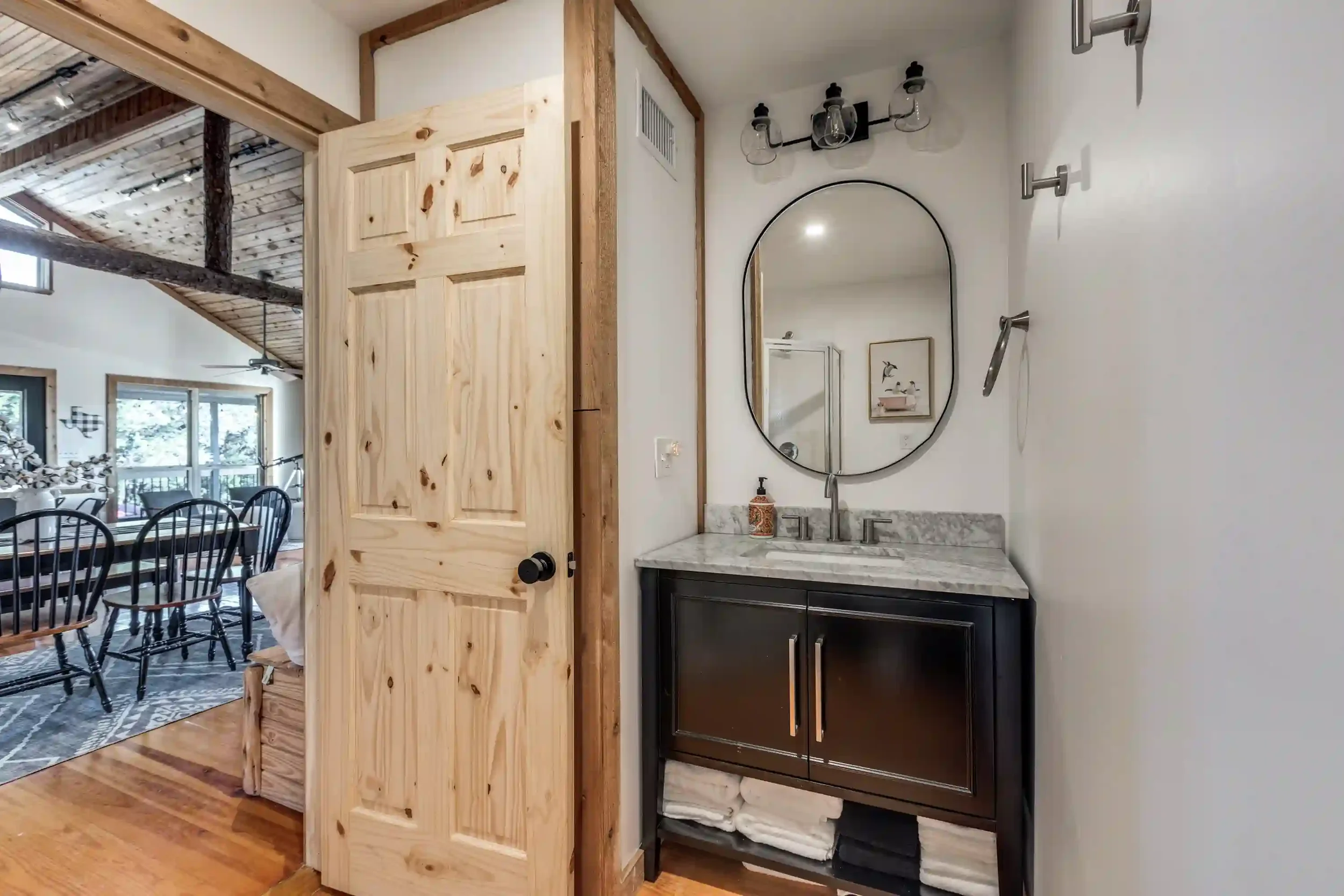 A bathroom with a granite countertop, a black vanity, and a large oval mirror. There is a soap dispenser on the counter, a wall-mounted light fixture above the mirror, and a partially open wooden door.