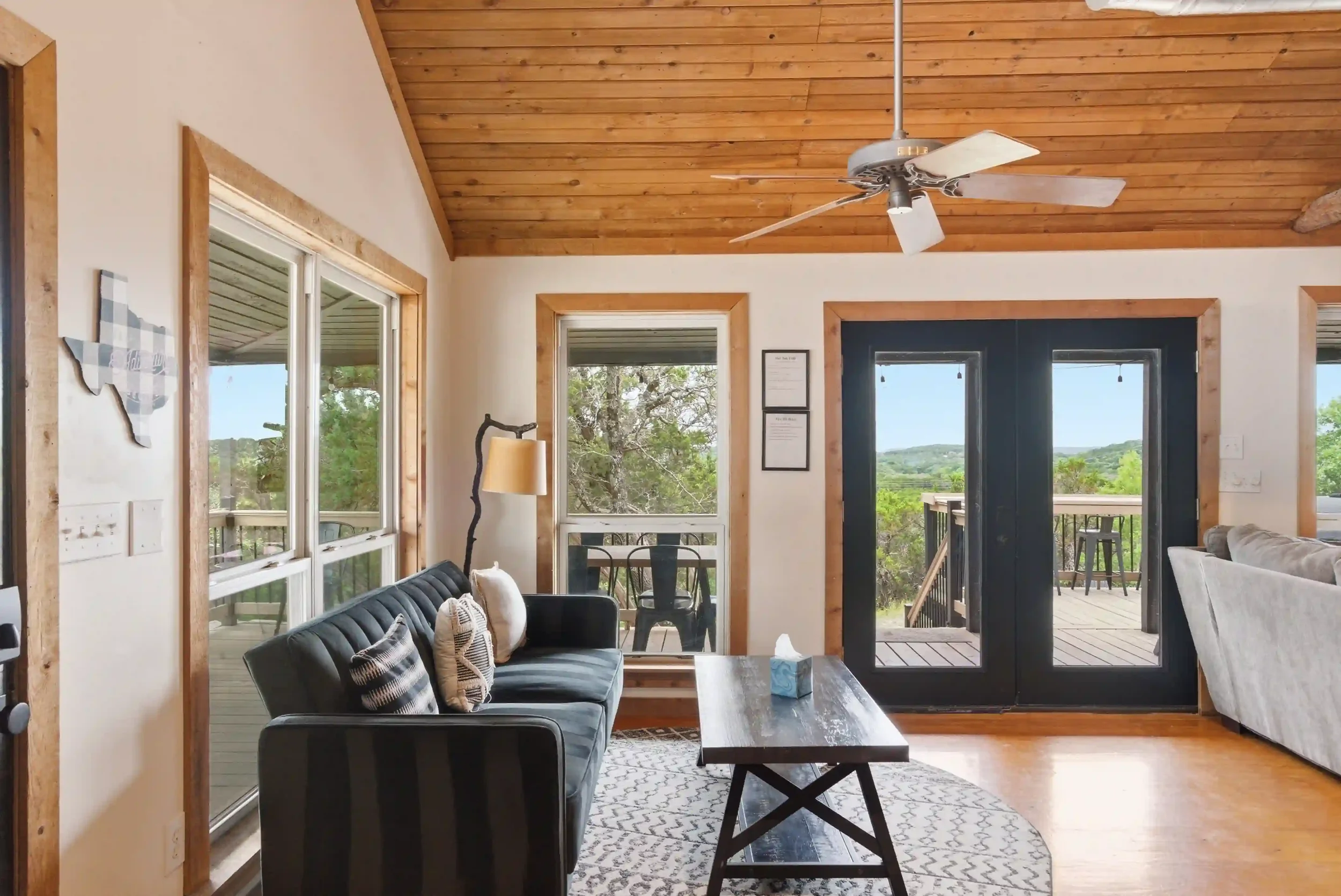 Sunlit living room with wooden ceiling and trim, black futon with pillows, gray sofa, wooden coffee table with blue tissue box, patterned rug, large windows showing trees outside, and a patio with outdoor furniture.