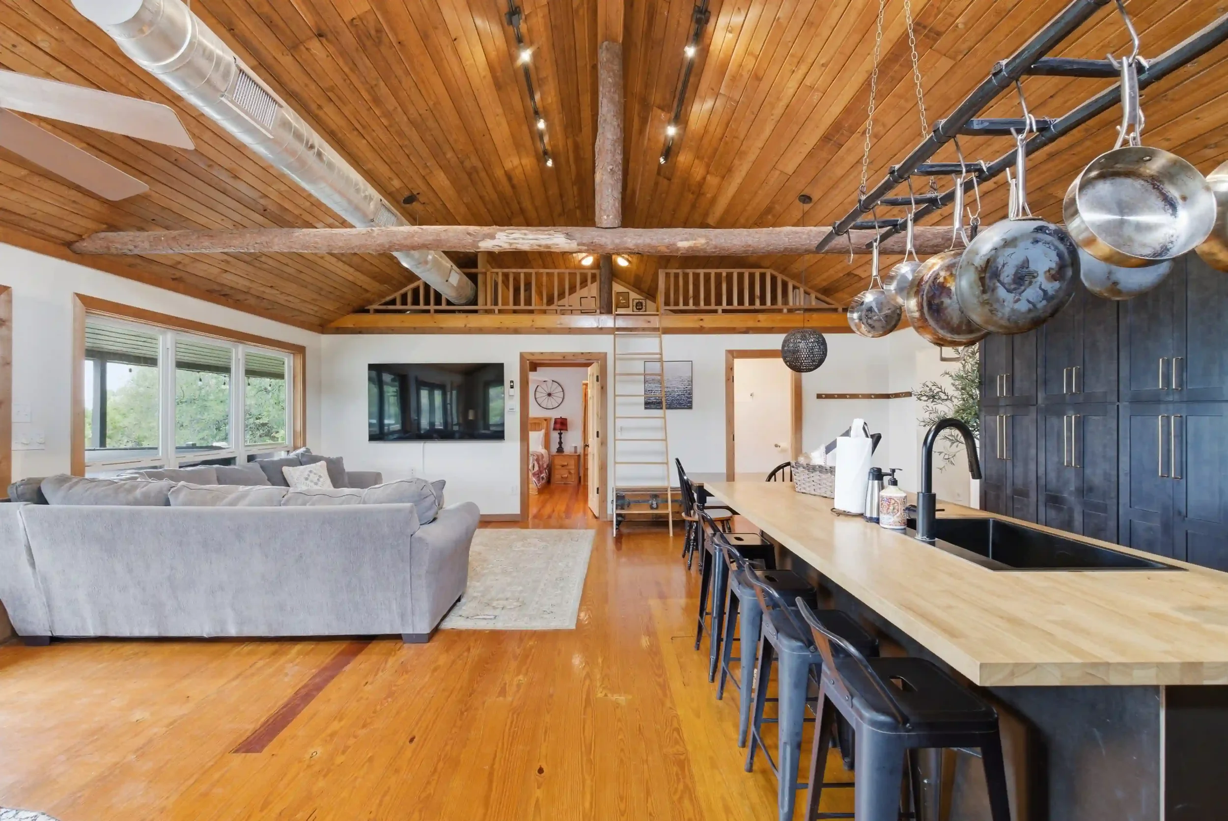 Open-concept living room with high wooden ceiling, large window, gray sectional sofa, wall-mounted TV, and kitchen island with black cabinets and hanging pots.