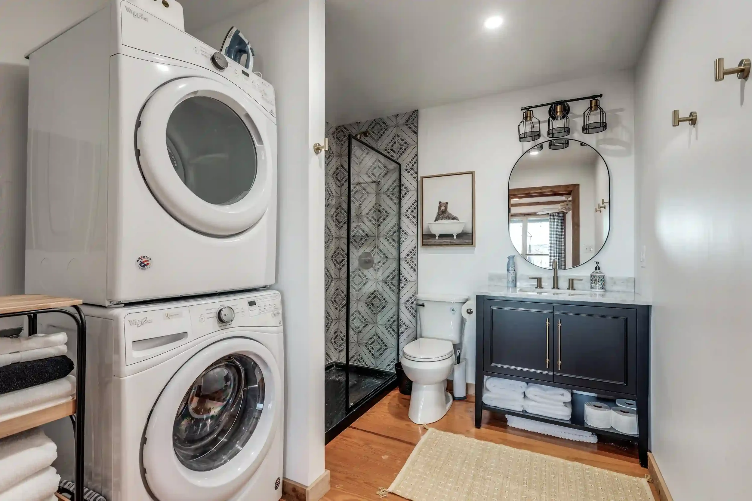 A modern bathroom with a stacked washer and dryer on the left, a glass shower enclosure with patterned tile, a white toilet, and a vanity with a mirror and lighting fixtures. There are shelves with towels and a wall art of a bear above the toilet.