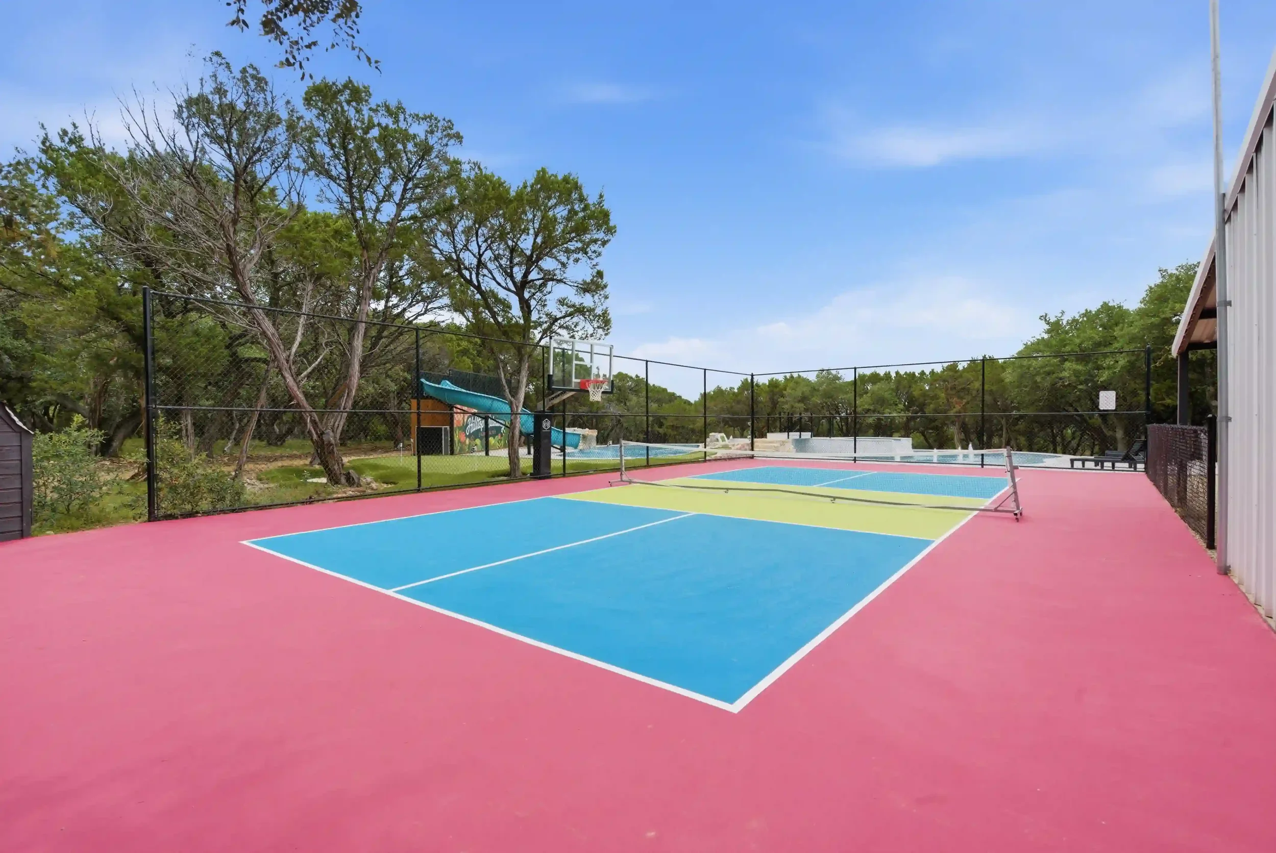 Colorful outdoor tennis or pickleball court with a basketball hoop and a small waterslide, surrounded by trees and a fence, under a blue sky.