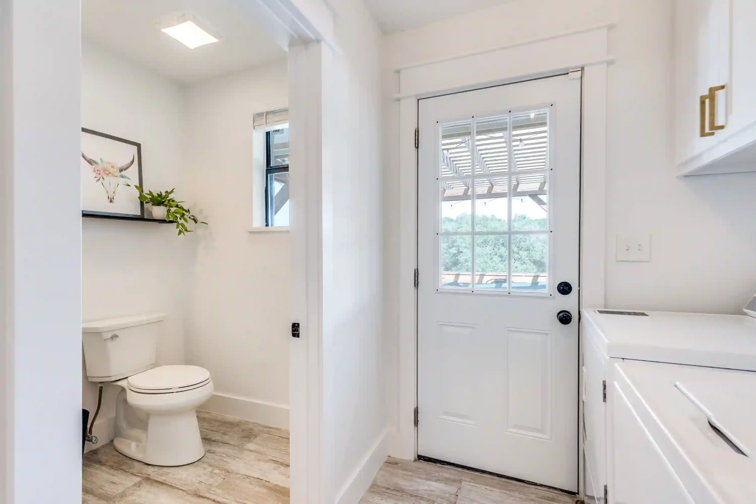Interior view of a bright, white laundry room with a door leading outside, a washing machine, and a small window with a decorative ledge and a potted plant. There's a visible toilet in an adjacent room.