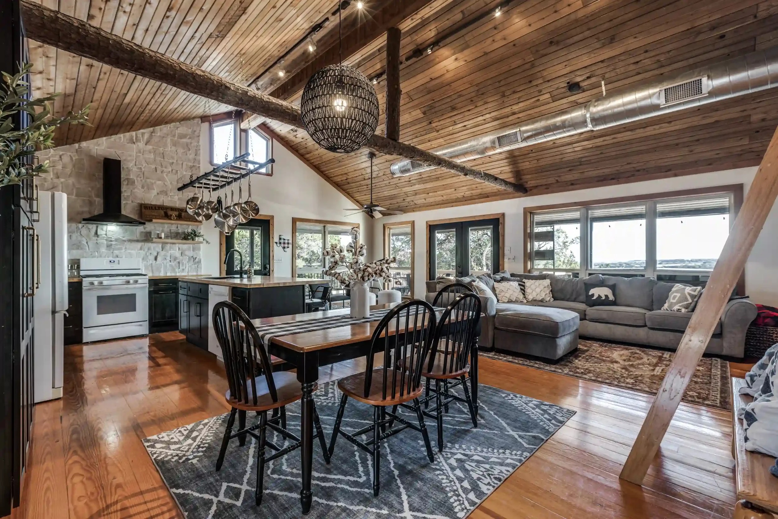 Open-concept living room and kitchen with wood vaulted ceiling, exposed beam, large windows, stone wall, and hardwood flooring.