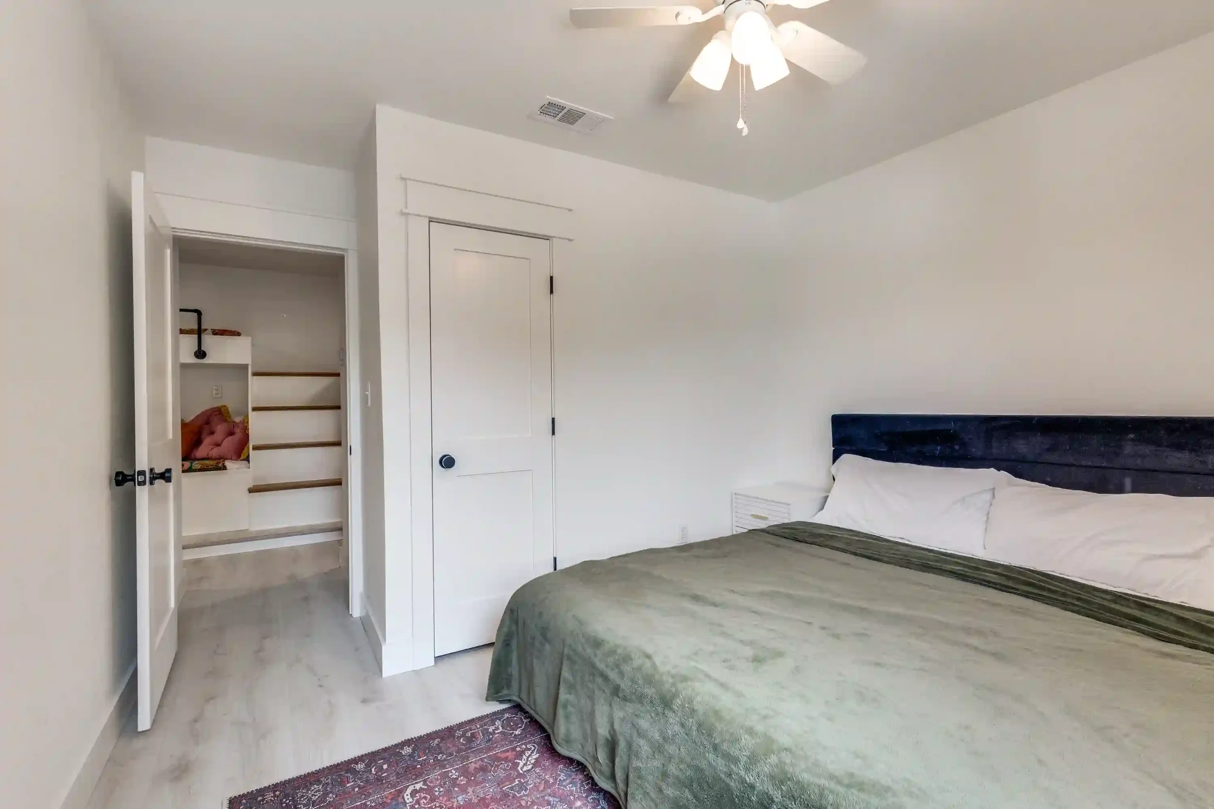 A bedroom with white walls, a white ceiling fan, and a bed with a green bedspread and white pillows. There is a small nightstand next to the bed, and a patterned rug on the light-colored wooden floor. An open closet with built-in shelves and a stairc