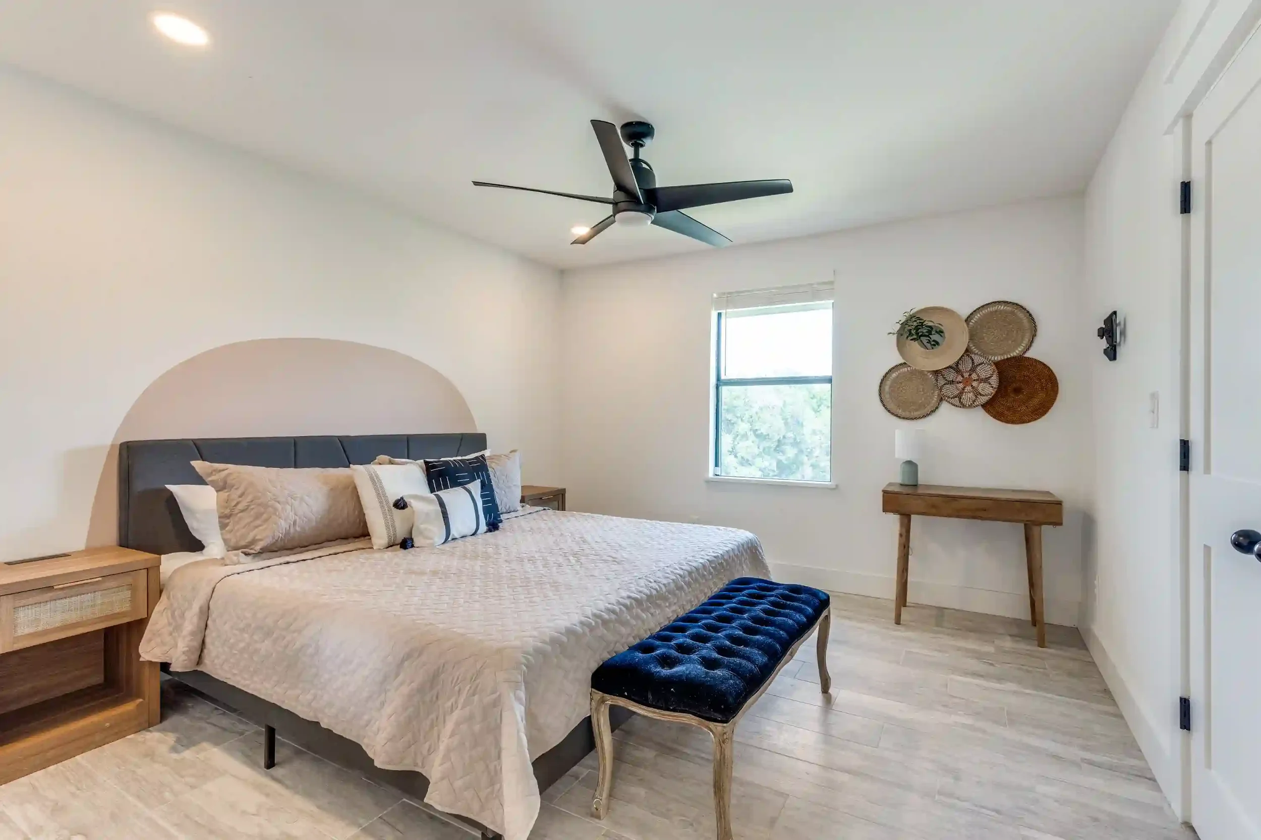 A bedroom with a bed, wooden nightstands, decorative pillows, a bench at the foot of the bed, a window with natural light, wall art made of woven baskets, a small table with a potted plant, and a black ceiling fan.