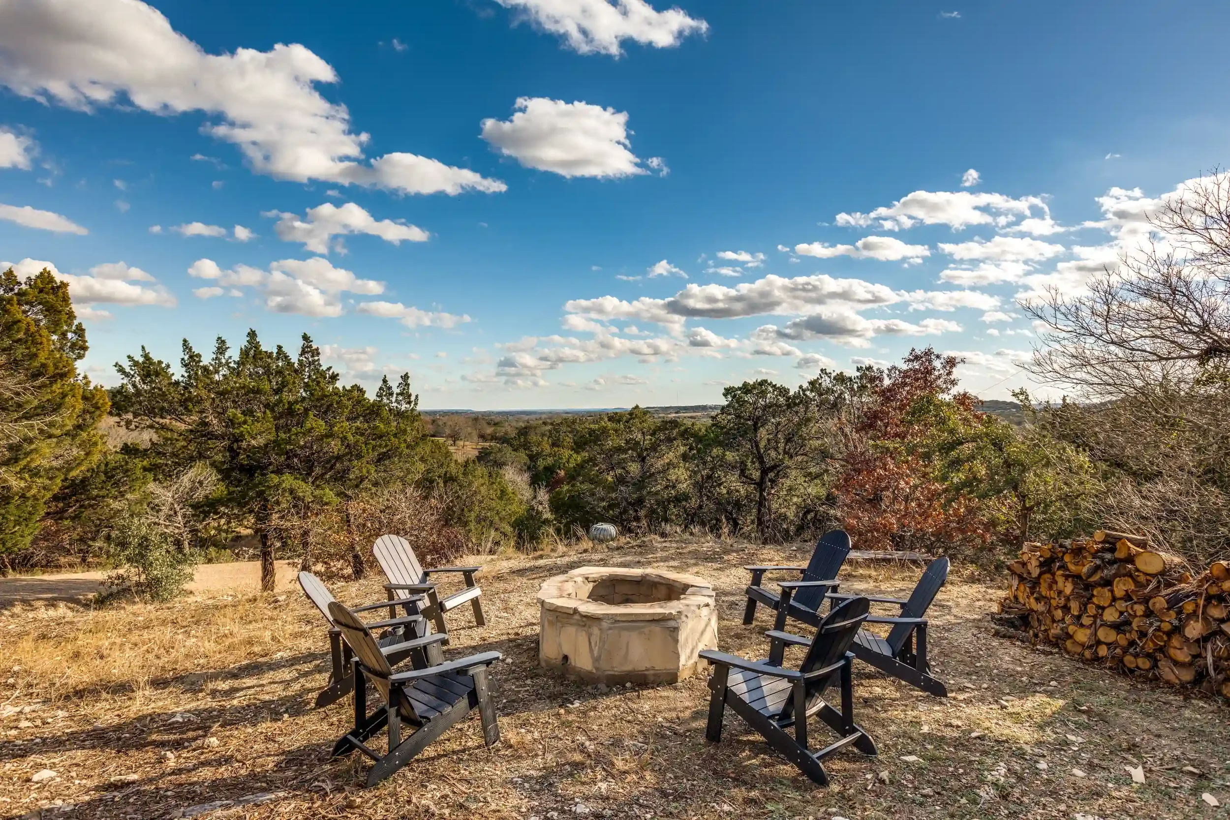 Outdoor fire pit area with six black Adirondack chairs arranged in a circle around a stone fire ring, surrounded by trees and a sunny blue sky with fluffy clouds.