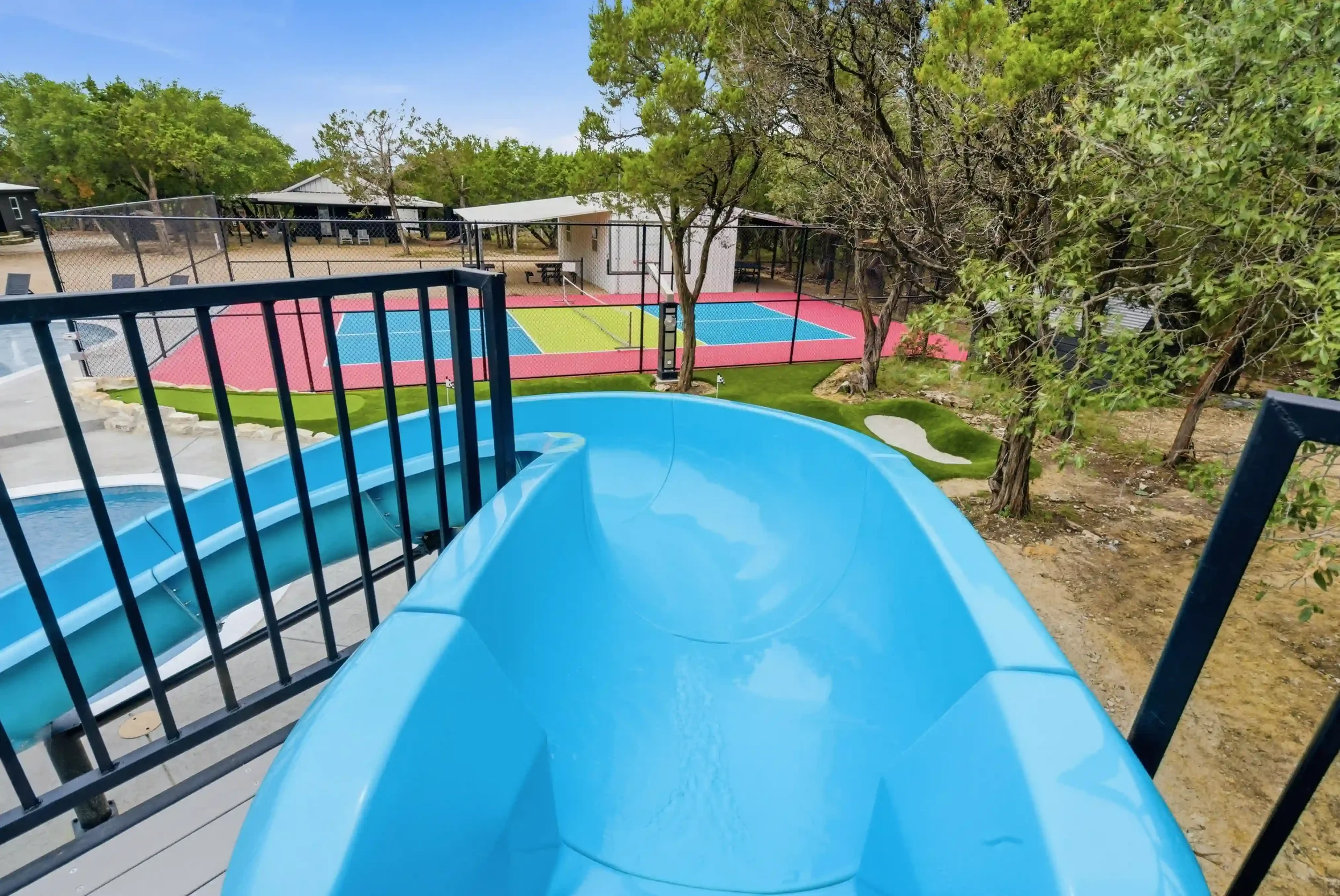A view from the top of a blue water slide overlooking a fenced tennis court with a pink basketball pickleball court