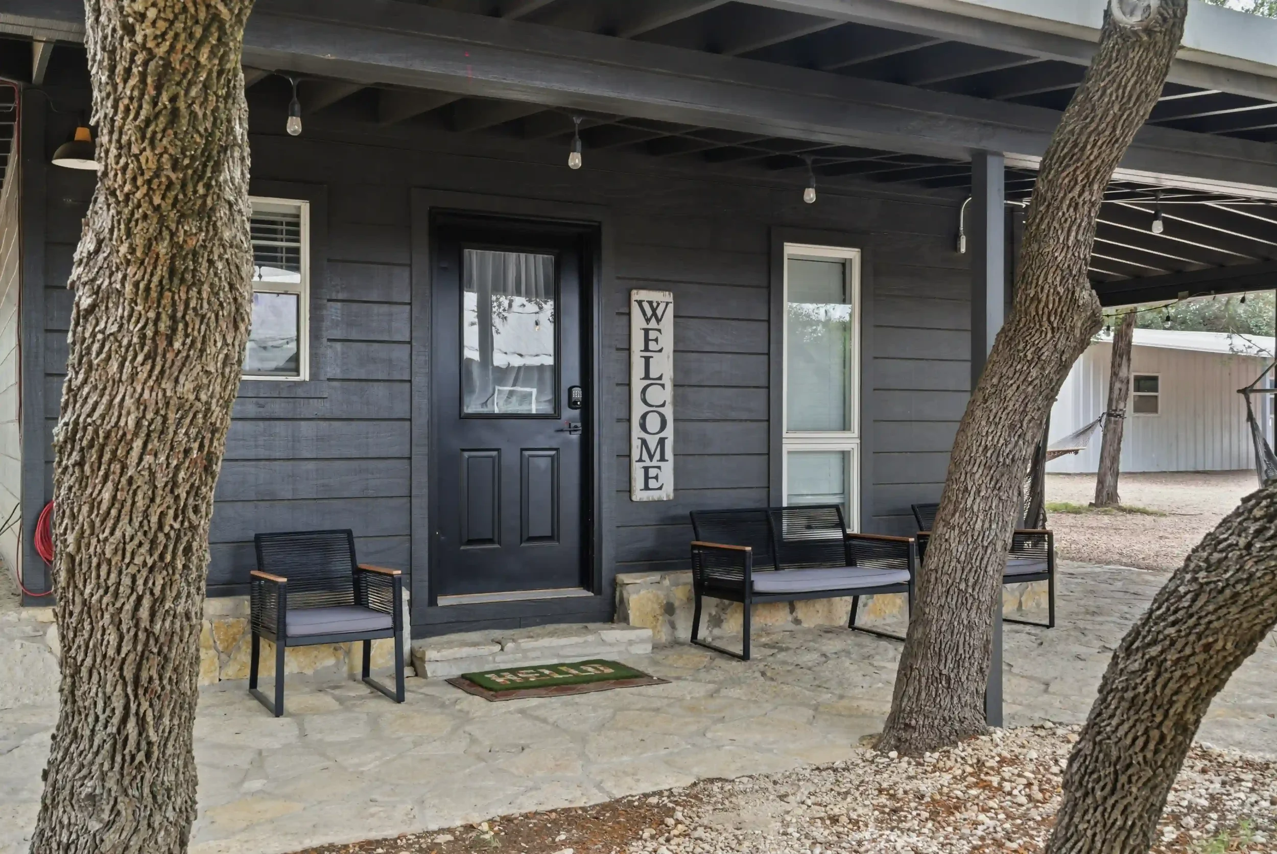 A gray house with a black front door, two windows, and a vertical welcome sign on the front porch. There are three black chairs on the porch and trees with rough bark in front of the house. String lights hang from the porch ceiling.