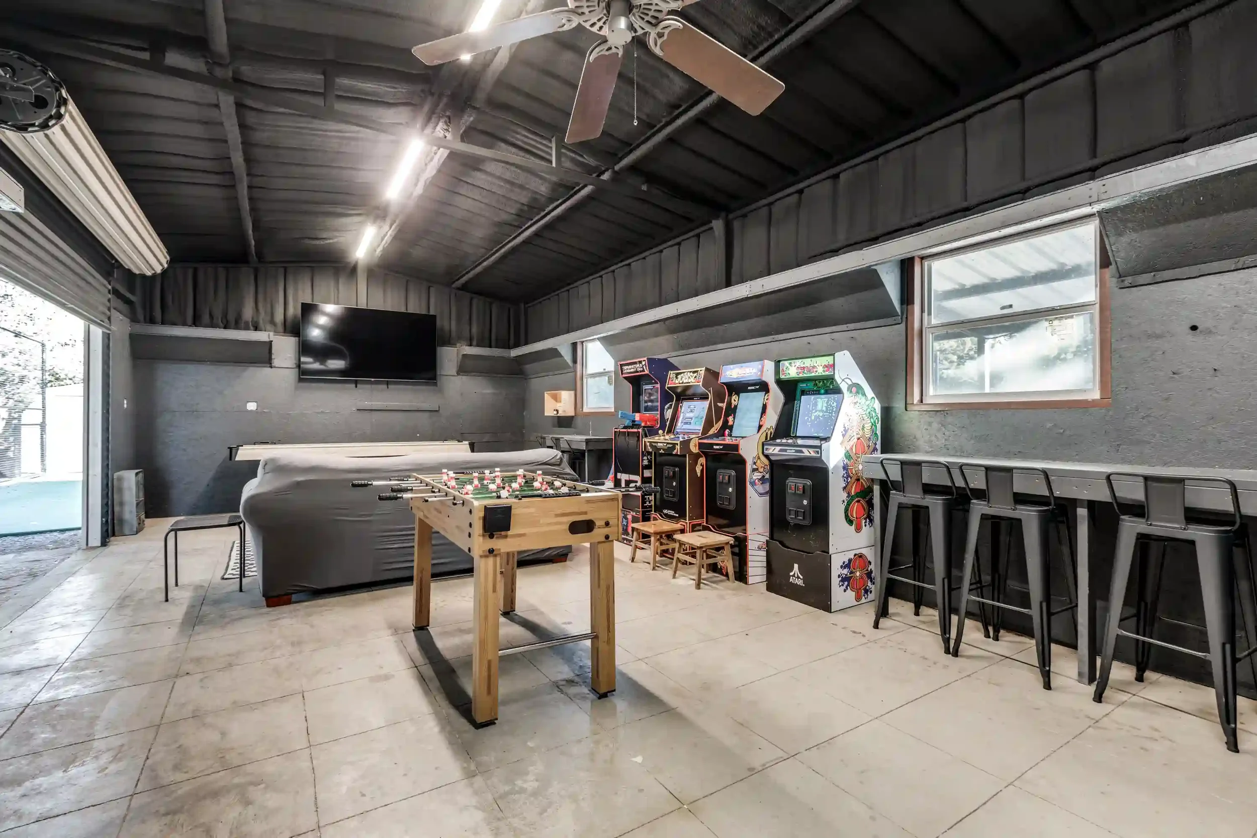 Indoor game room with arcade machines, a pool table, a foosball table, a television, and barstools near a window.