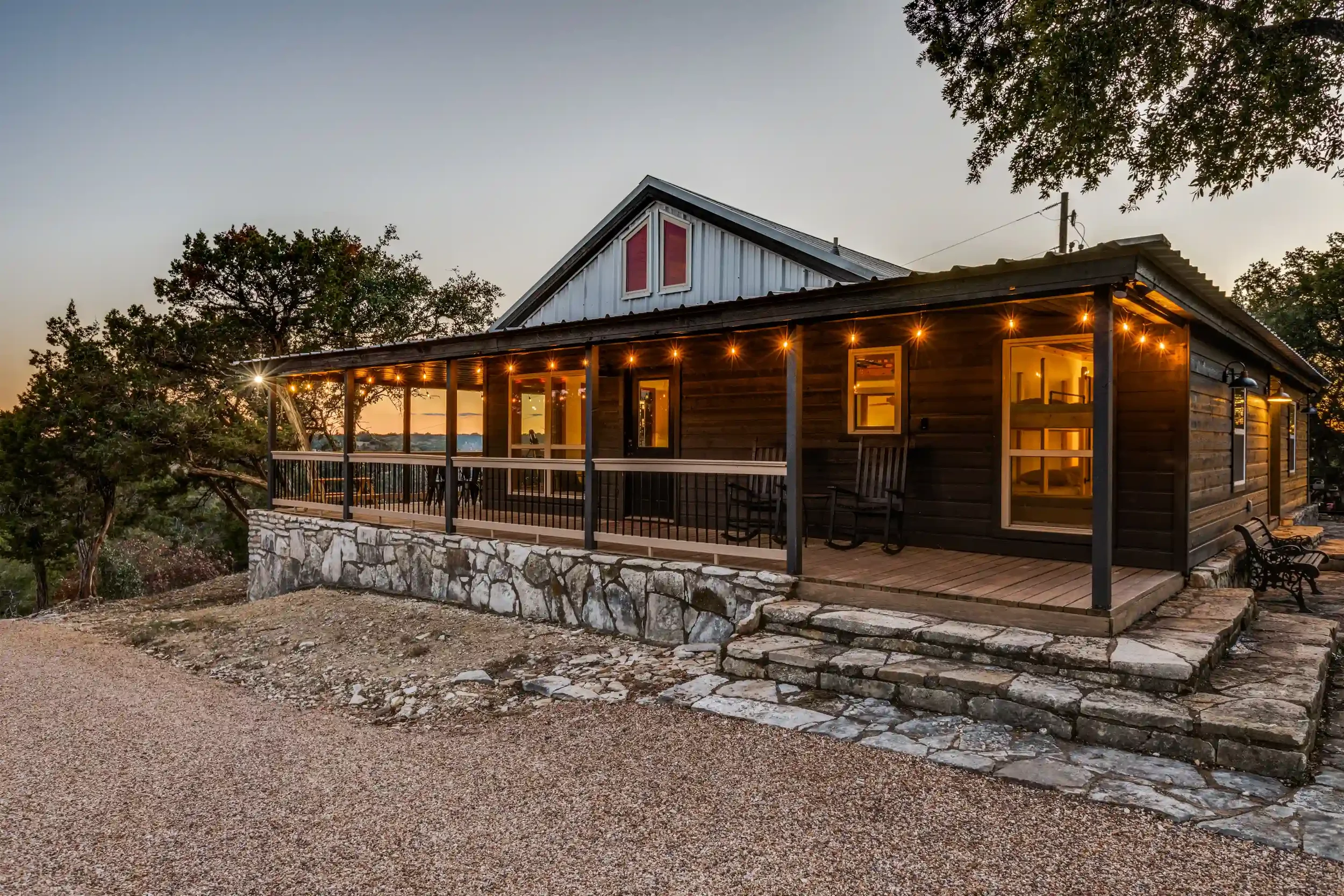 A wooden house with a balcony and string lights at dusk, surrounded by trees and a gravel pathway.