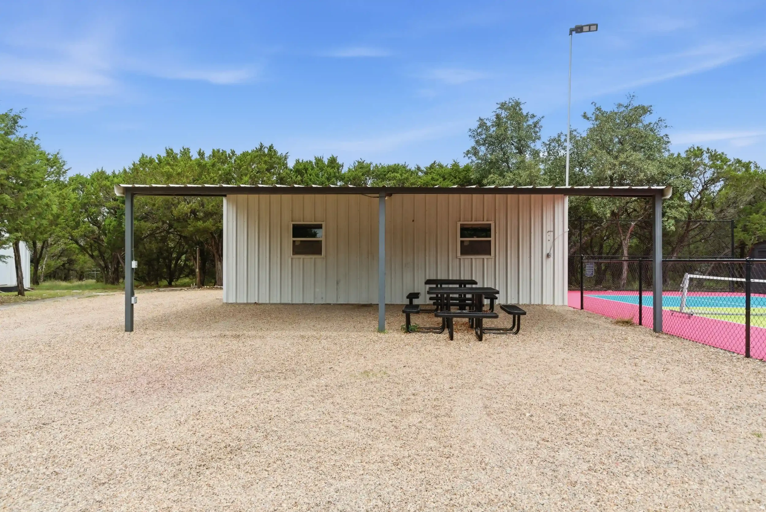 A small beige building with two small windows, surrounded by trees and a gravel ground, with a picnic table in front and a fenced colorful sports court to the right, under a clear blue sky.