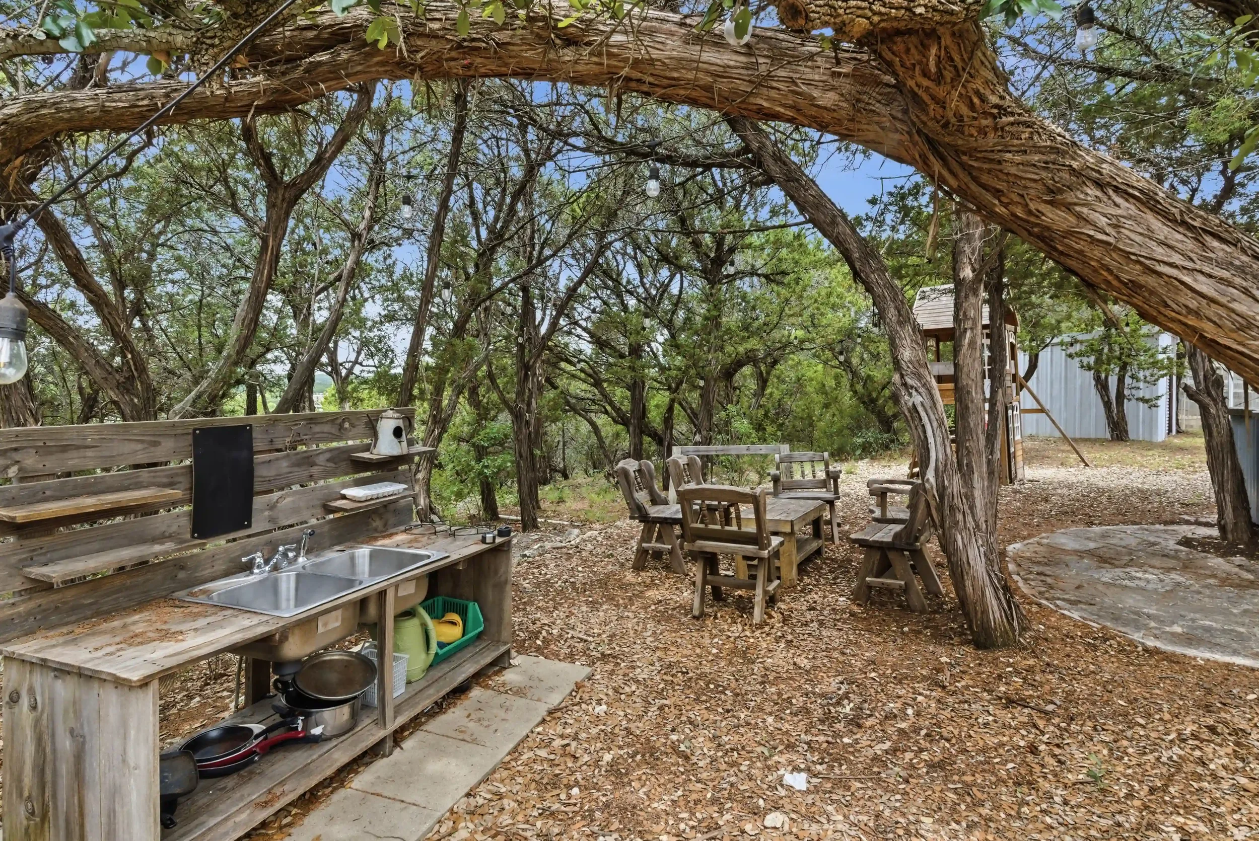 Outdoor backyard scene with rustic wooden furniture under large trees. There is a wooden outdoor sink with cleaning supplies, a sitting area with chairs, a treehouse, and string lights in a wooded yard.