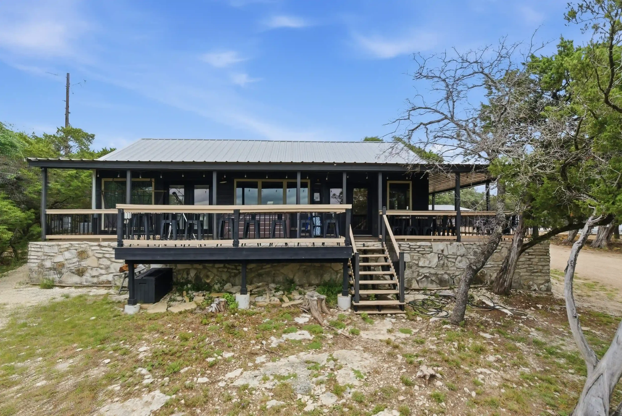 A house with a raised deck and stairs, made of black siding and a metal roof, surrounded by trees and grass under a blue sky.