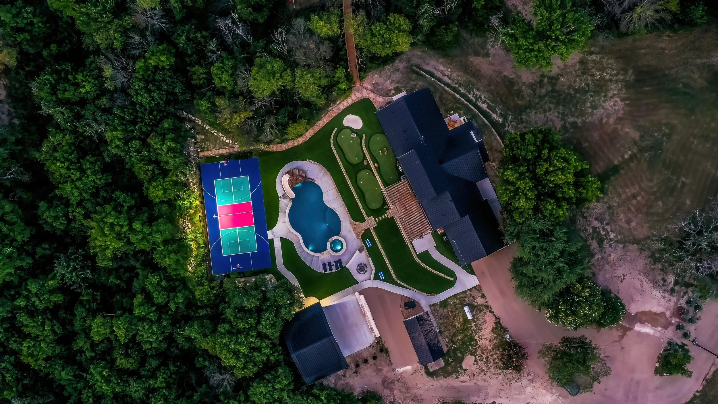 Aerial view of a backyard with a pool, a basketball court, a house, and surrounding trees.