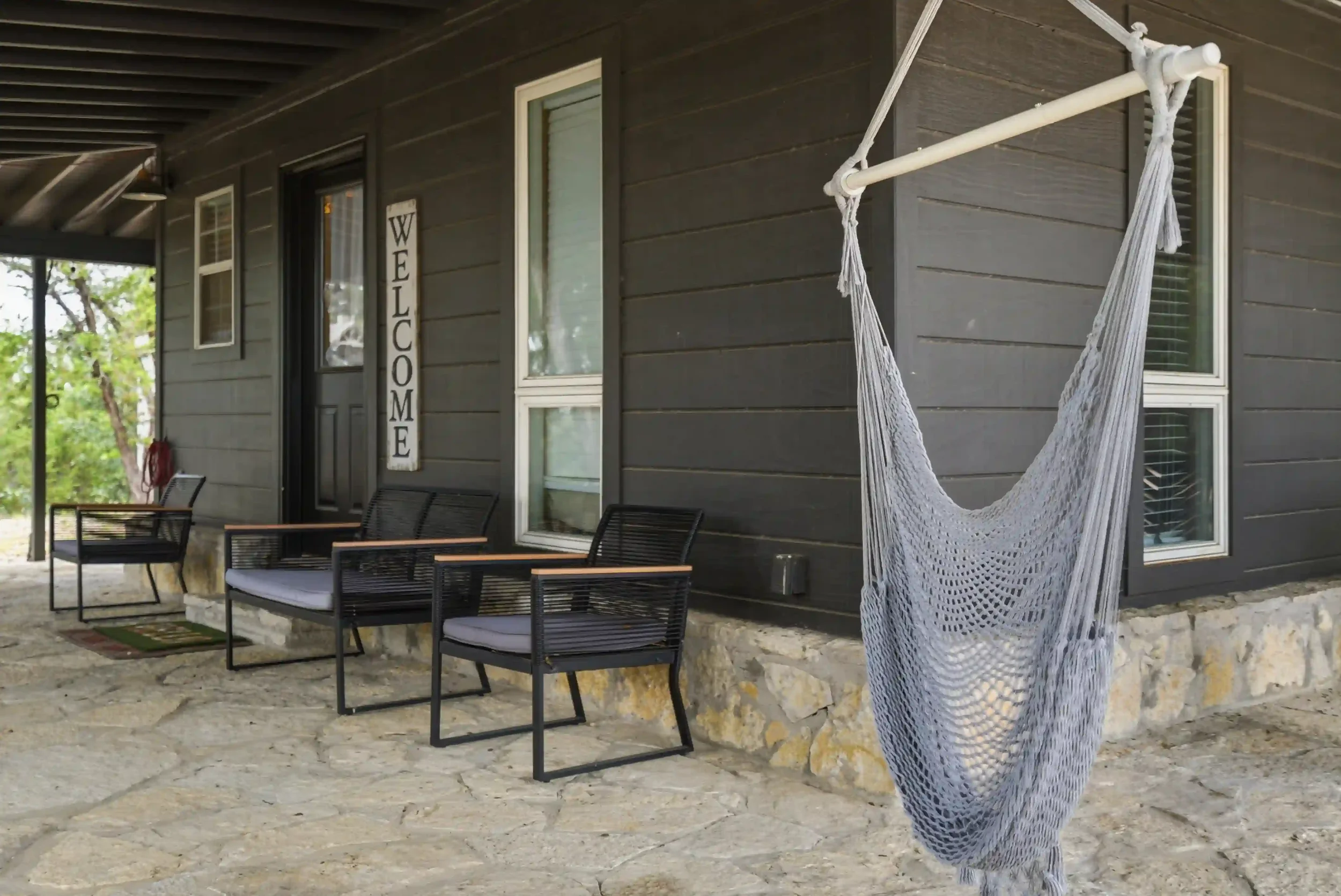 A porch with black chairs and a hammock hanging from the ceiling, in front of a dark gray house with a vertical 
