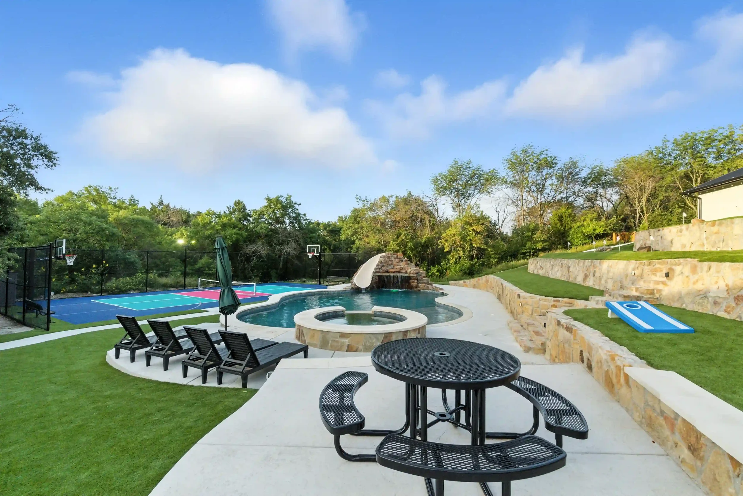 An outdoor backyard with a pool, hot tub, and a basketball court surrounded by trees under a partly cloudy sky.