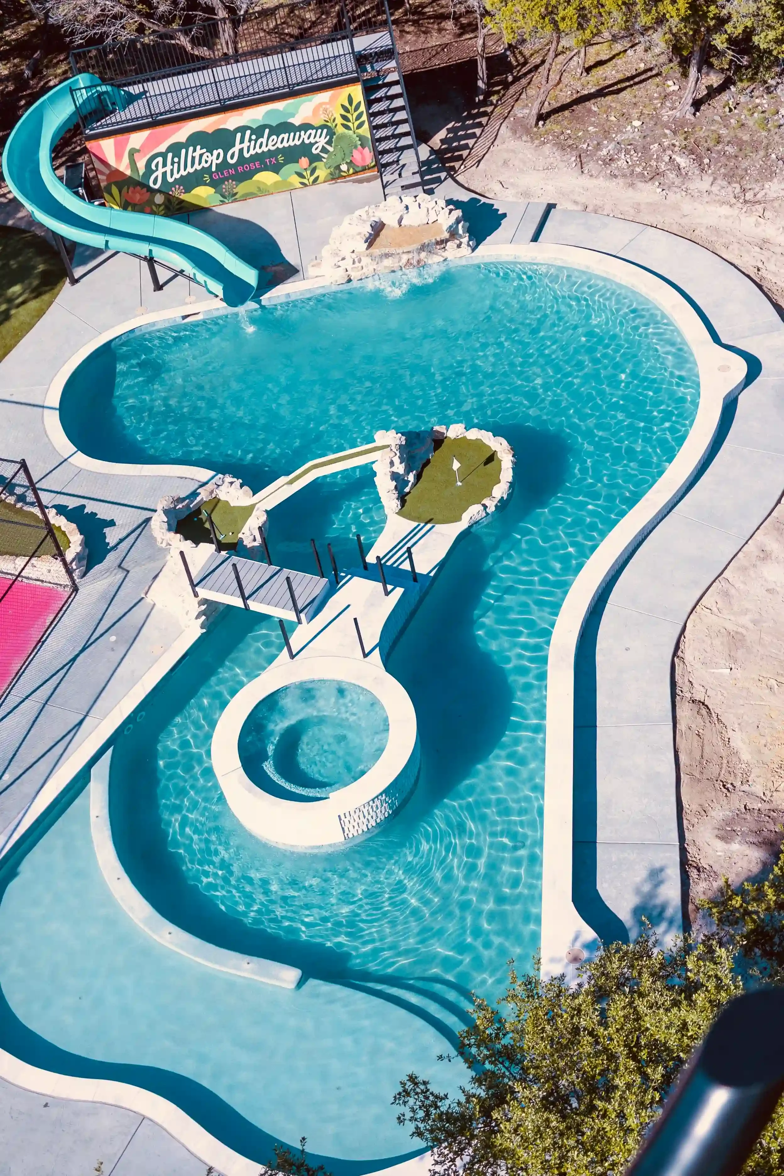 Aerial view of a swimming pool with a water slide and a small circular hot tub, surrounded by a concrete deck and some greenery, with a sign in the background reading "Hilltop Hideaway, Glen Rose, TX."