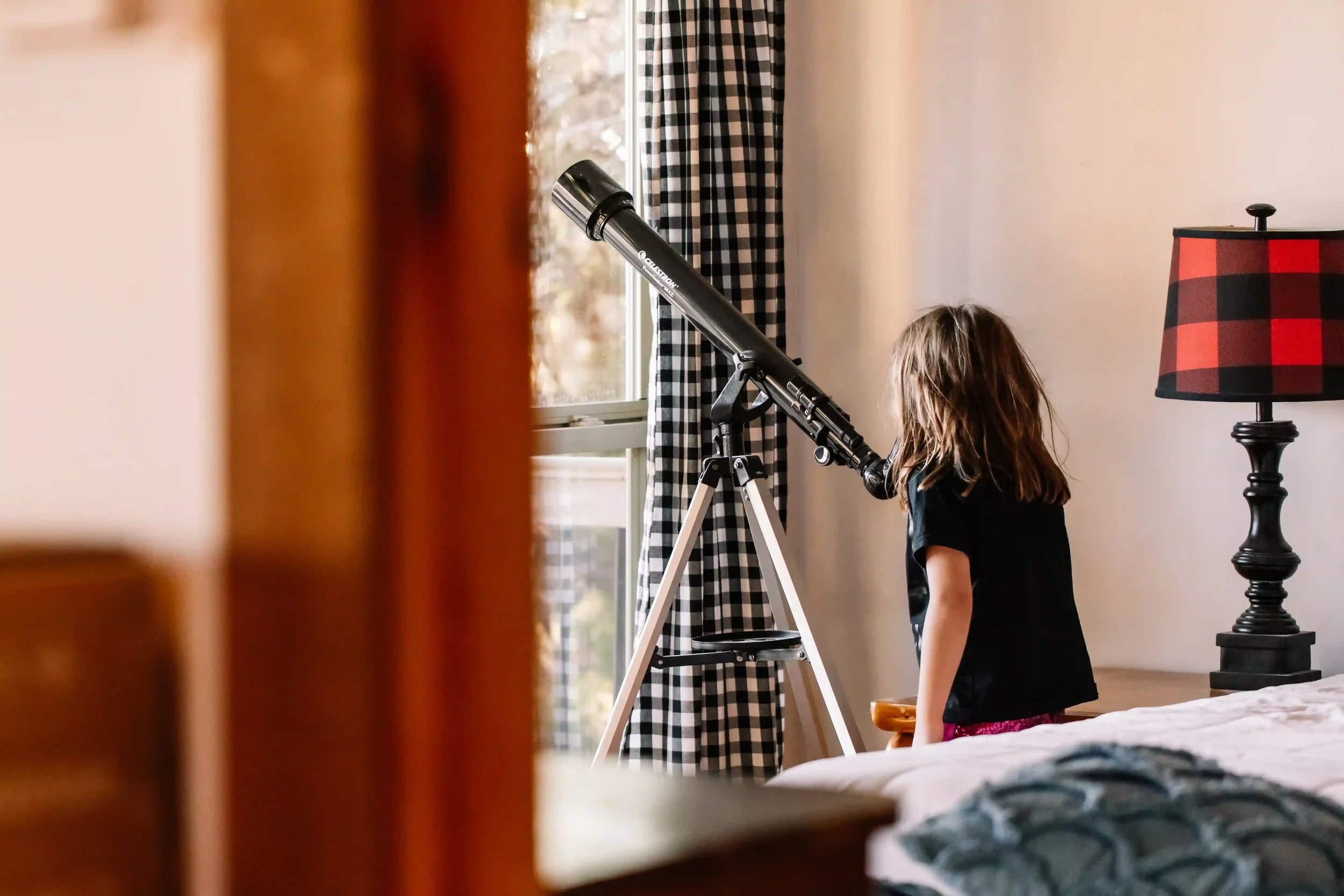 A young girl with long hair looking through a telescope near a window with black and white checkered curtains in a bedroom, with a lamp with a red and black checkered shade on a black stand nearby.