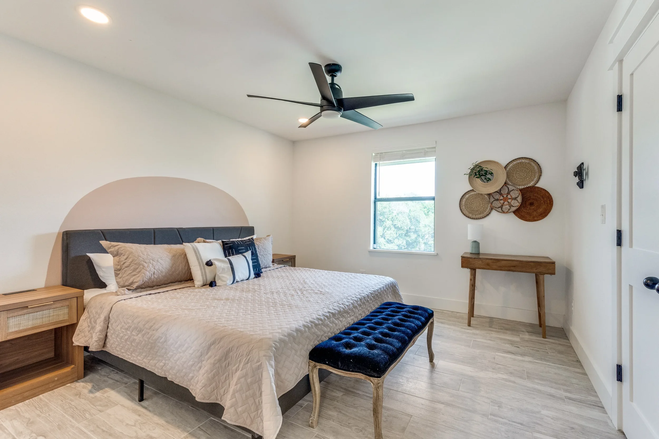 A bedroom with a bed, bedside tables, wicker wall decor, window, ceiling fan, and a bench at the foot of the bed.