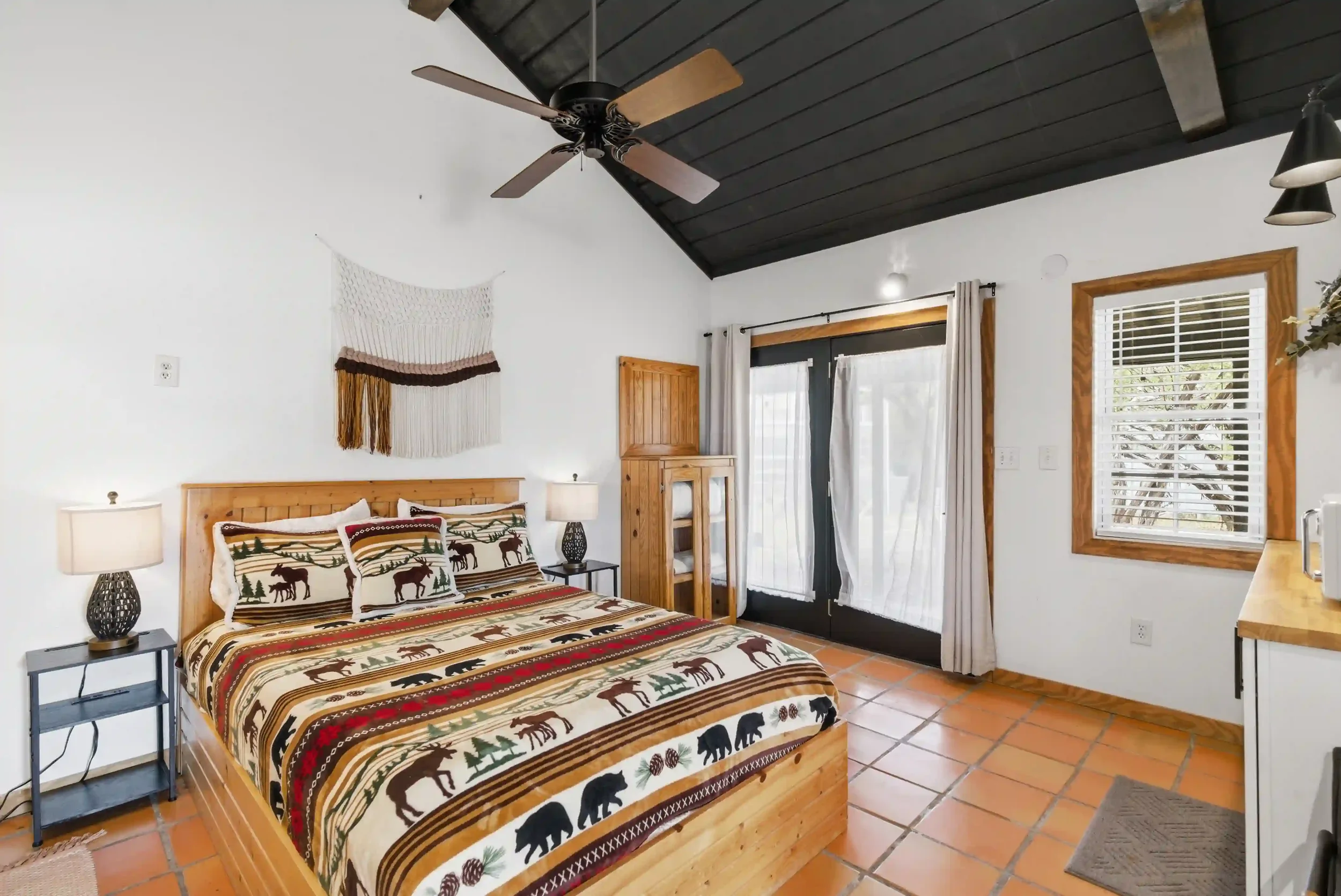 Bedroom with wooden bed frame and bedding featuring animals and nature patterns, two nightstands with lamps, wooden storage cabinet, windows with white curtains, black door with curtains, ceiling fan, and tiled floor.