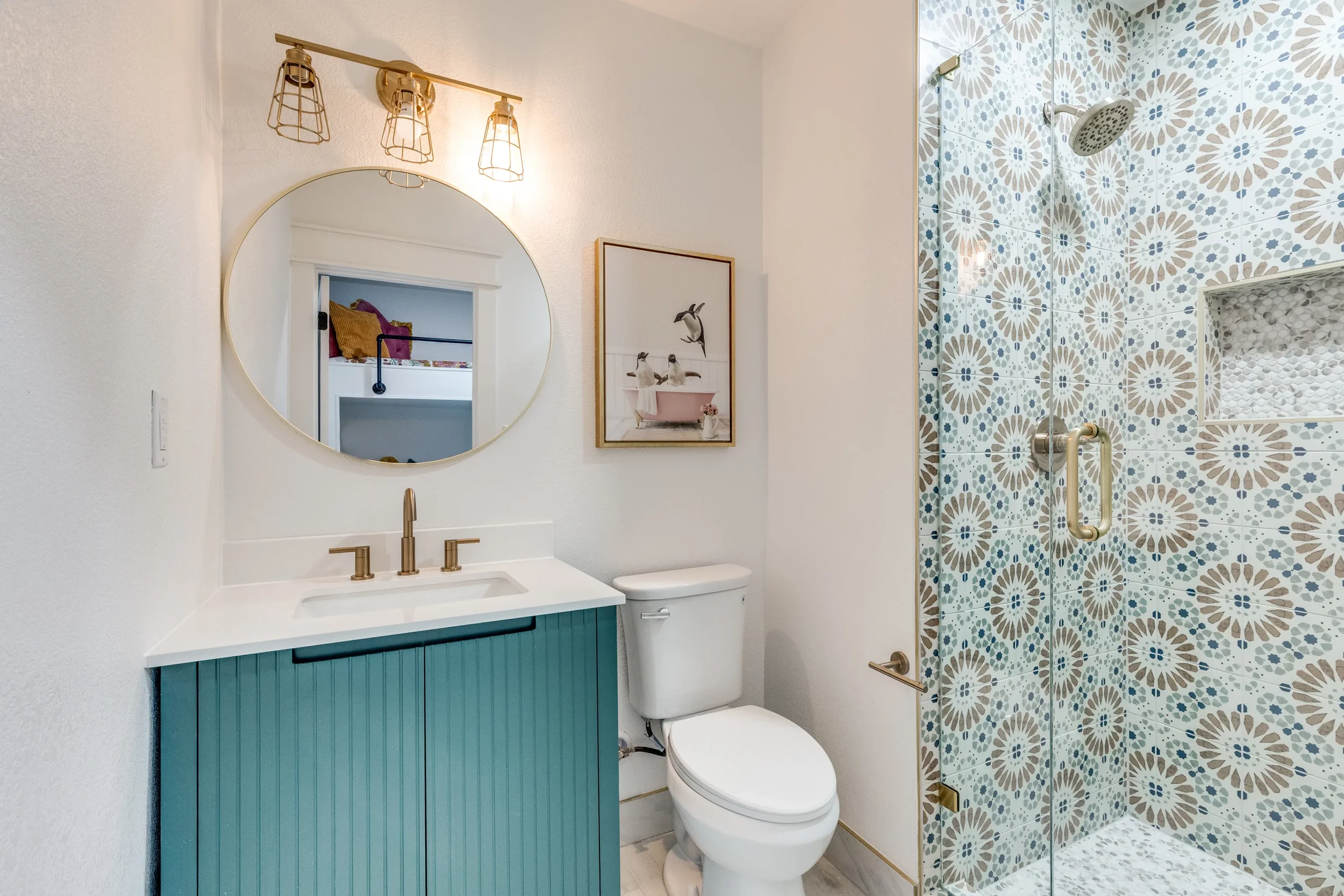Modern bathroom with a teal vanity, round mirror, gold fixtures, a white toilet, and a glass shower with patterned tiles in shades of blue, beige, and white.