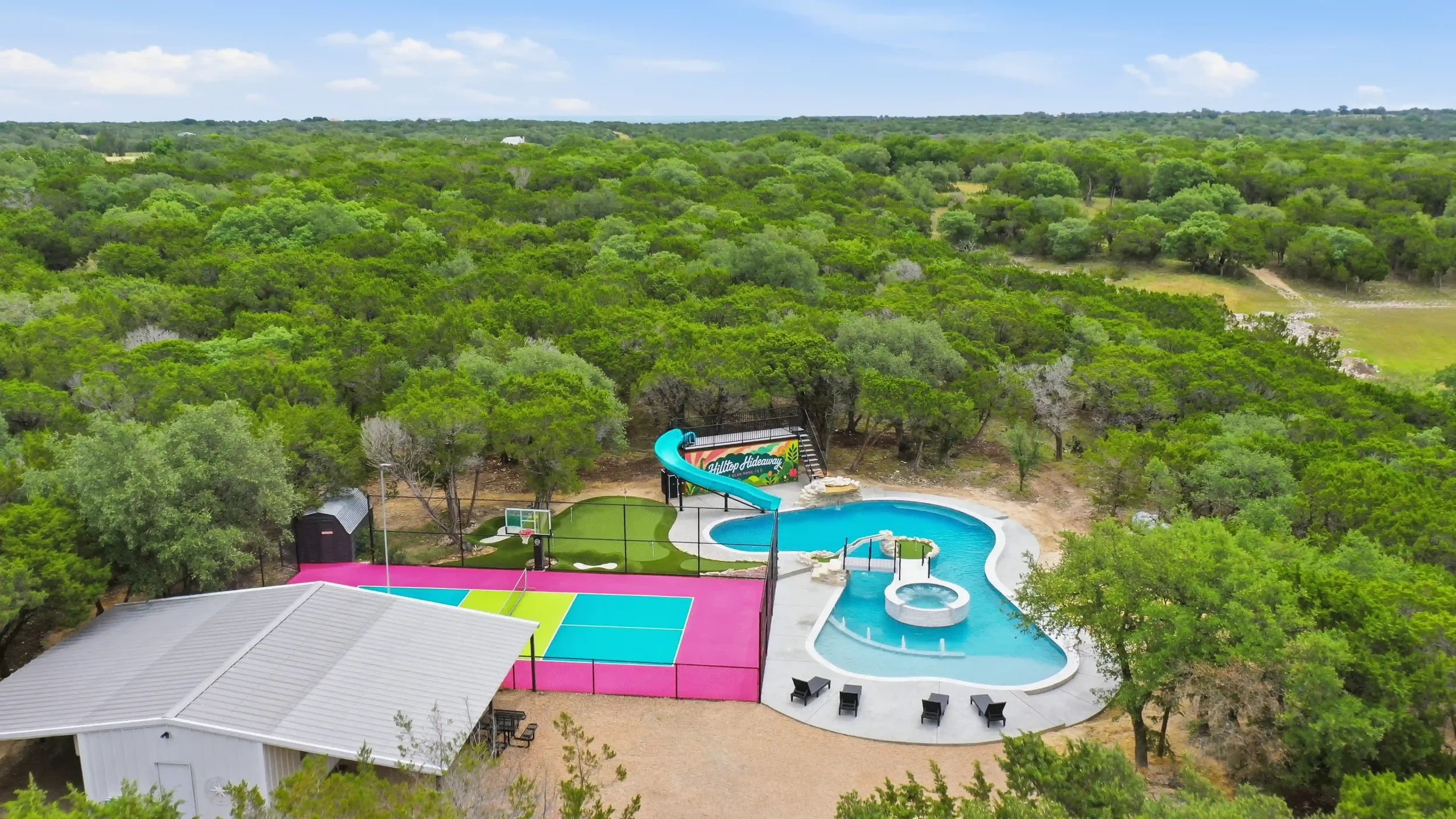A backyard with a pool, waterslide, basketball hoop, and a volleyball court, surrounded by trees and open green landscape under a blue sky with clouds.