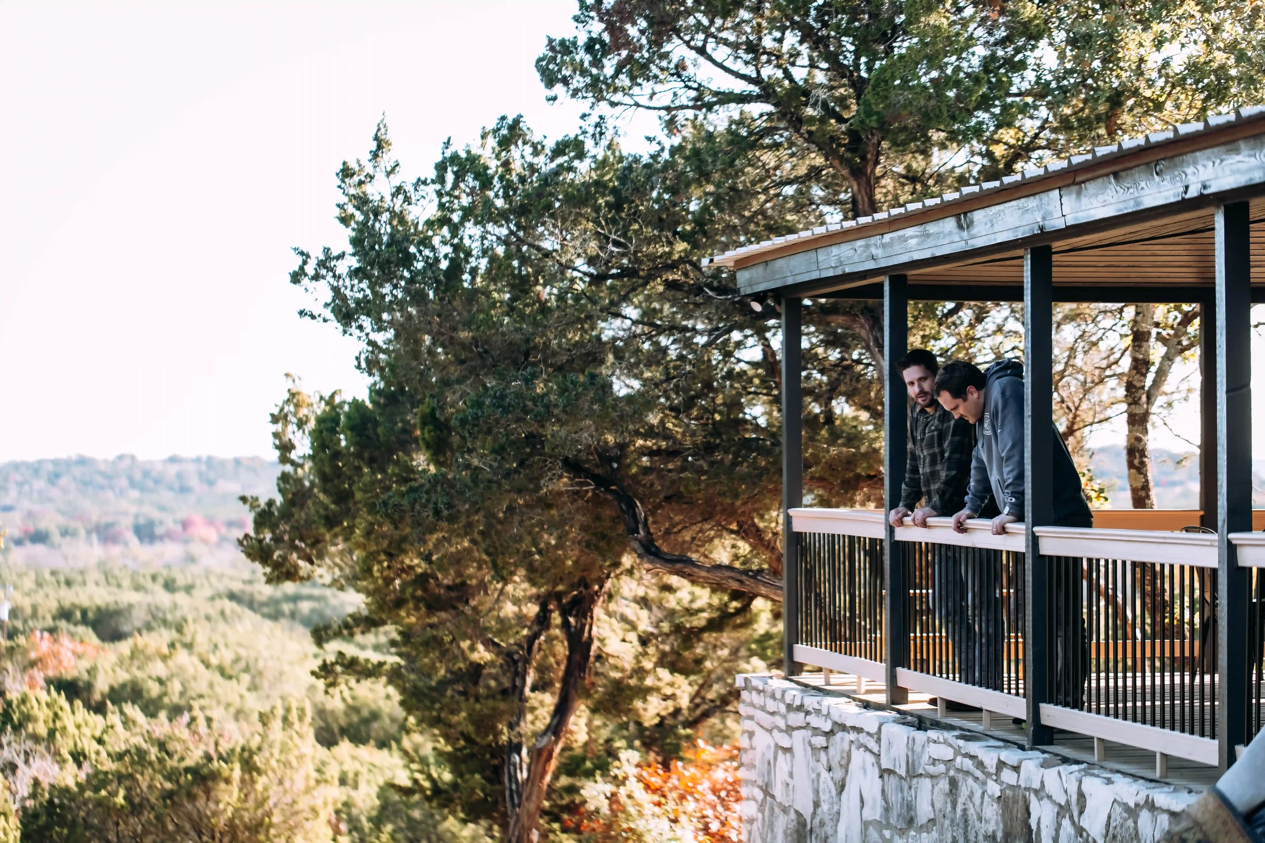 Two men leaning over a balcony railing on a house with a wooden roof, overlooking a landscape of trees and distant hills.