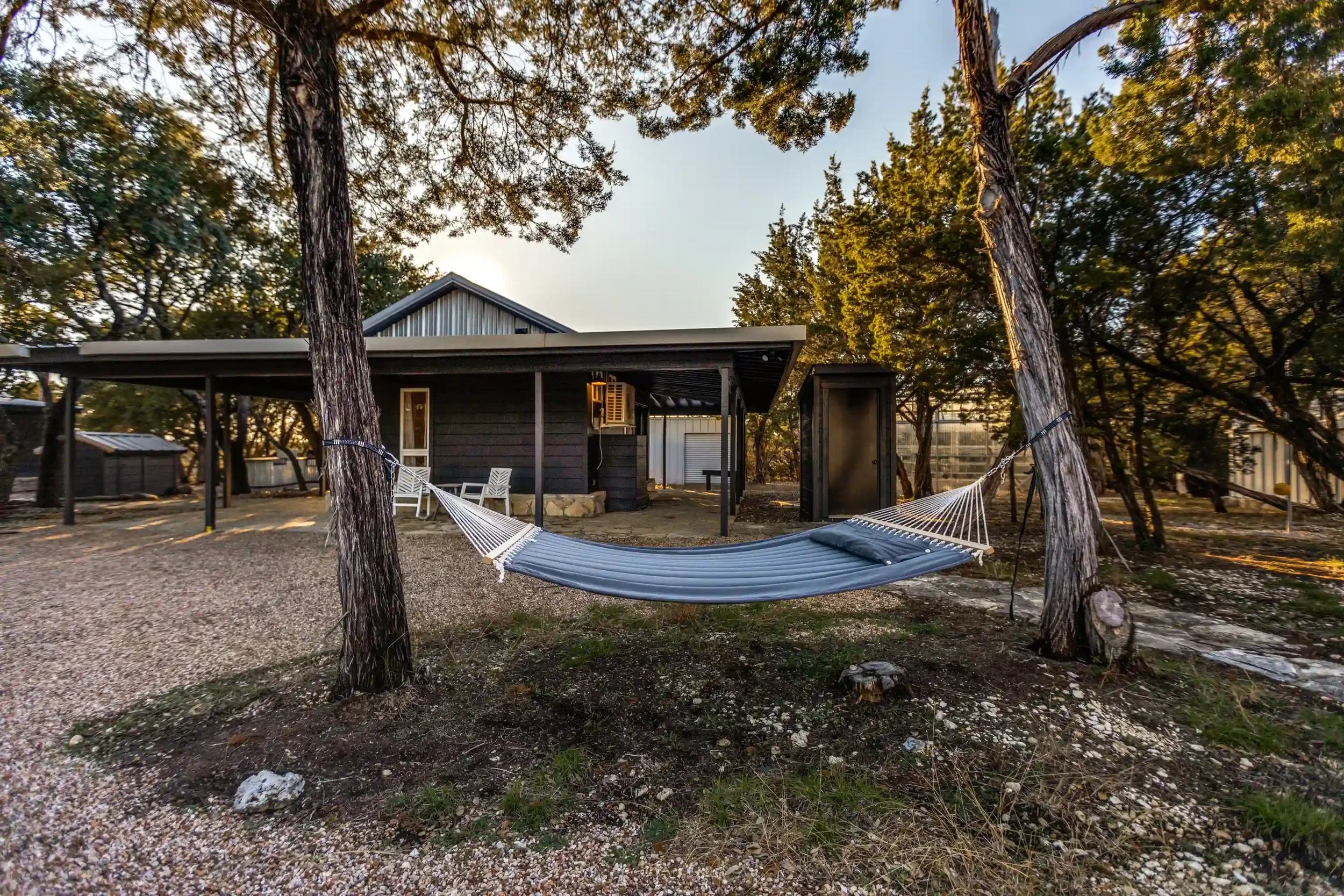 Outdoor scene with two tall trees, a hammock between them, and a small building with a covered porch in the background during sunset.