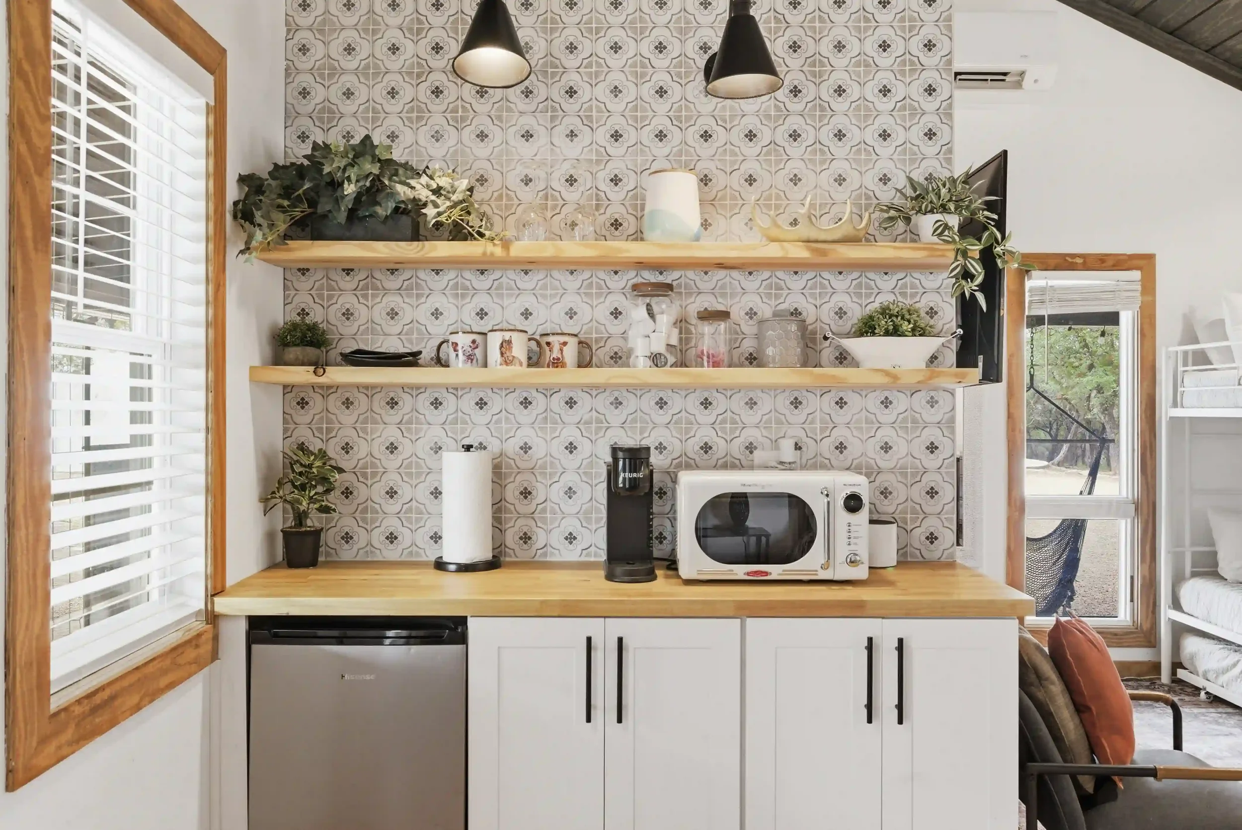 Kitchen with open wooden shelves, patterned tile backsplash, black pendant lights, potted plants, and small appliances including a microwave and coffee maker.