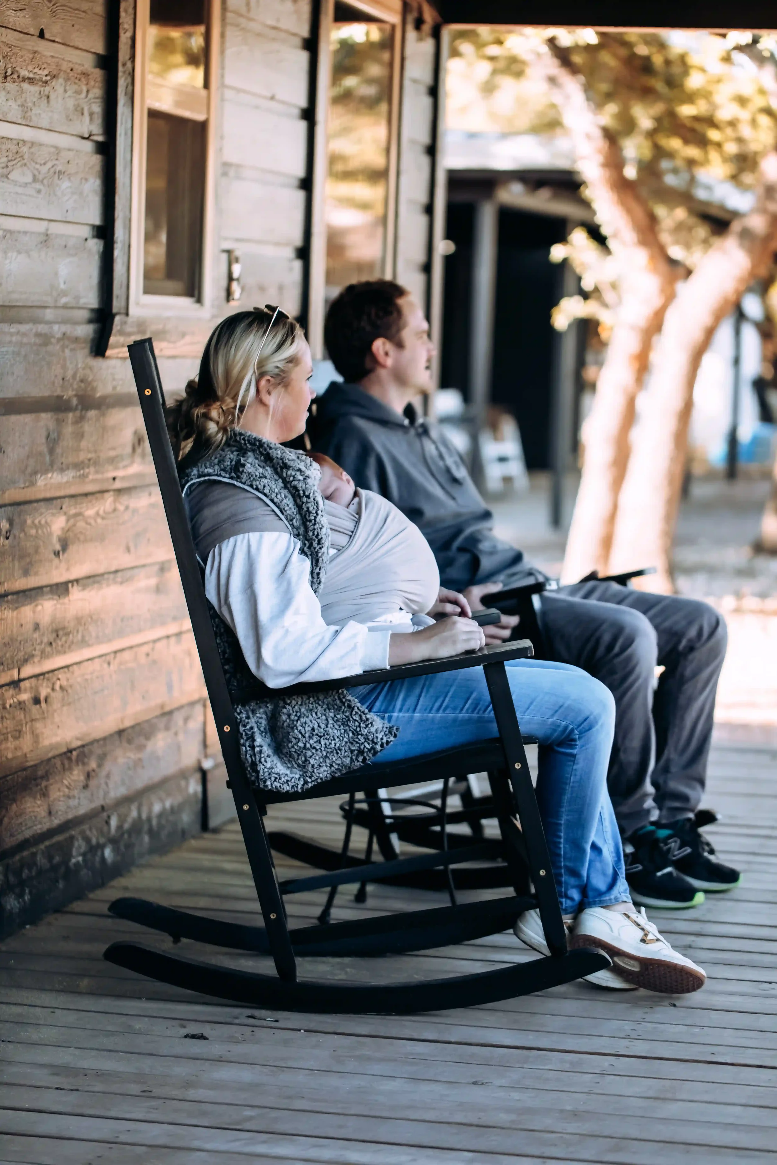 A woman and a man sit on wooden rocking chairs outside a rustic wooden building, with trees in the background. The woman is holding a sleeping baby, and they are all relaxed and facing the same direction.